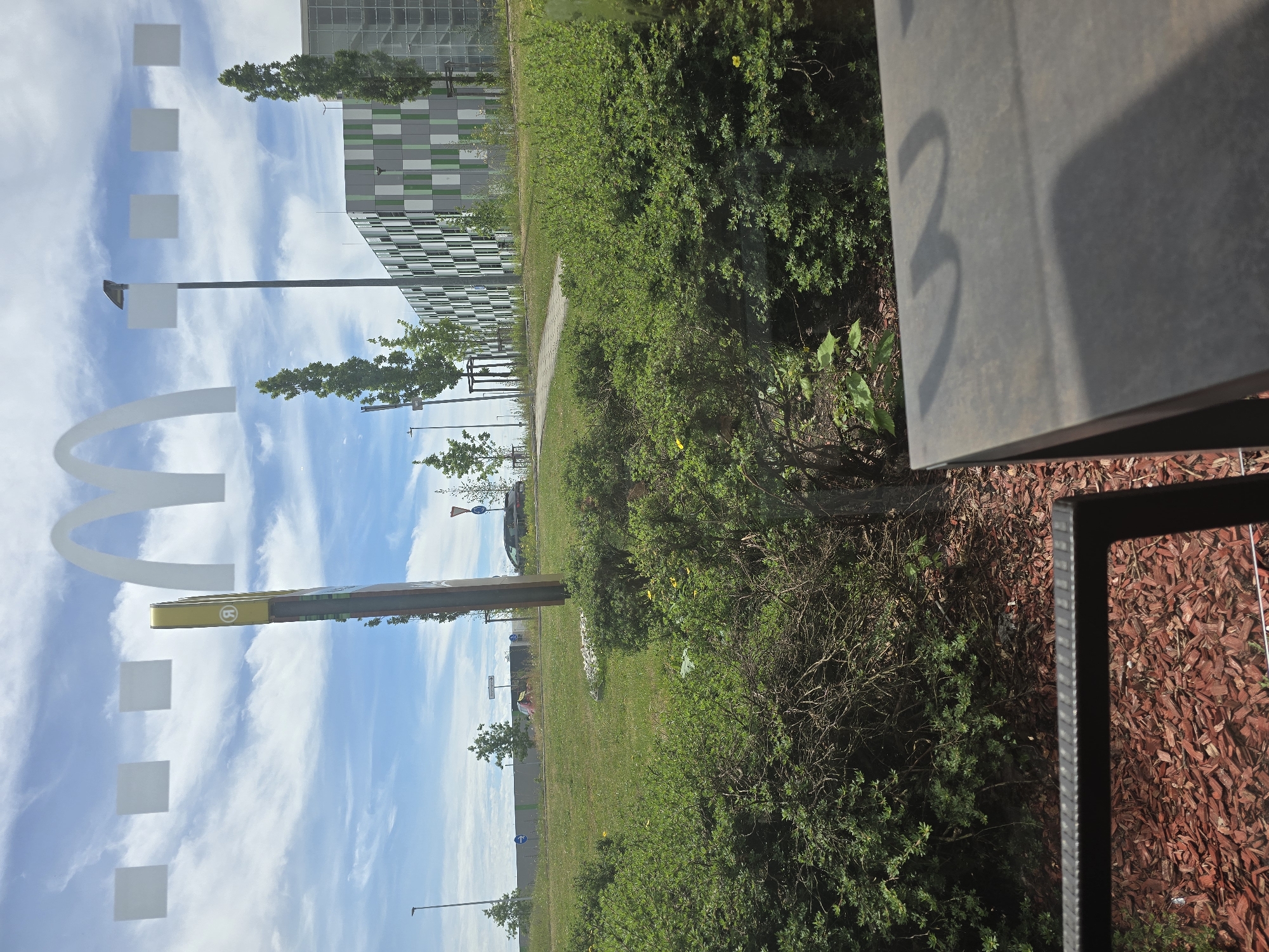 The image presents a detailed outdoor scene viewed through a window, indicated by the prominent reflection of McDonald's golden arches and white square accents across the top of the frame. The observation point is from within a building, likely a McDonald's restaurant in the city of Erding, Germany.
In the immediate foreground, a dark metal frame, possibly part of a chair, is visible on the bottom left. To its right, a dark, flat tabletop or counter extends, casting dark shadows from objects not fully visible. Just outside the window, a bed of reddish-brown wood chips (mulch) covers the ground.
Beyond the mulch, dense green bushes, interspersed with small yellow flowers and some dry branches, form a natural barrier. A narrow paved path can be seen winding through the foliage, leading towards the middle ground. The bushes transition into a wide grassy lawn area.
Mid-ground elements include several tall, slender evergreen trees and smaller deciduous trees scattered across the lawn. On the left, a tall black lamppost stands, with another shorter lamppost further to its left. A prominent, tall yellow pylon sign, characteristic of McDonald's, is positioned on the left side of the grassy area. It features a circular white emblem at its top, displaying a stylized letter 'R' that appears as 'Я'.
Behind the lawn, a road is visible, with a light-colored vehicle and a dark vehicle in the distance. Road signs include two blue circular signs with white right-pointing arrows and a red and white triangular yield sign on the right. A low, gray concrete barrier or wall runs along the far side of the road.
The background is dominated by a large, modern, multi-story building on the right, featuring a distinctive facade composed of alternating rectangular panels in dark grey, light grey, and light green. The sky above is partly cloudy with significant patches of blue, indicating a bright, clear day. No people are visible anywhere in the scene.