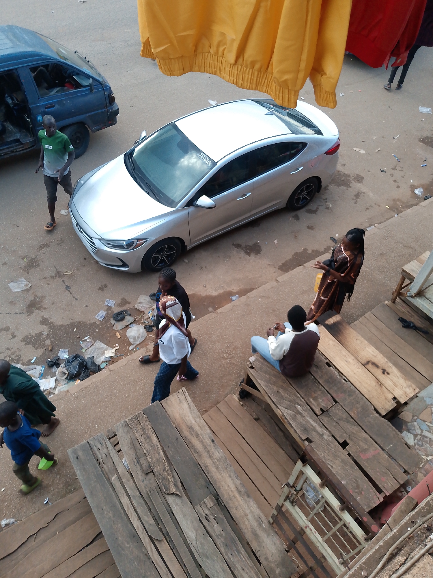 A street scene in Bukuru, Nigeria, captures a moment of daily life with a mix of vehicles, people, and litter. In the foreground, wooden planks form a makeshift path or structure. A young child in a blue shirt and green pants walks across the scene, carrying a green object. To the left, a person in a dark green garment walks barefoot. Further up the street, a silver sedan is parked, and to its left, a blue van is also parked. A man wearing a green t-shirt and dark shorts stands near the van. A woman in a patterned brown dress stands facing a seated man in a white and brown shirt and blue jeans, on wooden steps. Another woman in a white shirt and patterned pants walks with a younger person. A red cloth and a yellow cloth hang overhead, partially obscuring the view. The street itself is paved and shows signs of wear and tear, with scattered litter.