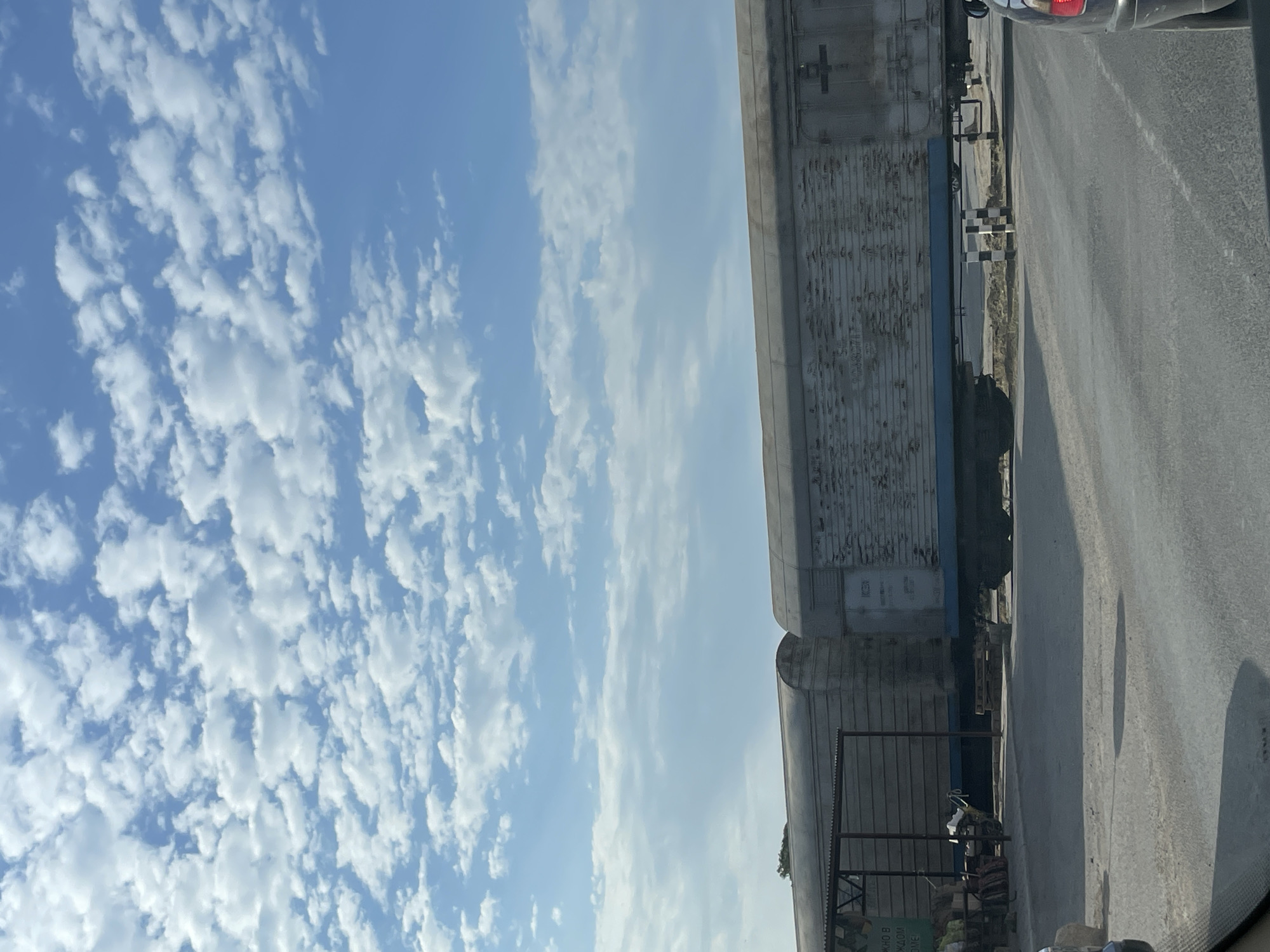 The image depicts a railway crossing in Aktau, Kazakhstan, with two train carriages visible. The carriages appear to be old and weathered, with peeling paint and rust. The sky above is a clear blue with scattered white clouds. A paved road runs in front of the train tracks, and a black car is partially visible in the foreground on the right. A small, open-sided shelter with a metal roof is located to the left of the train carriages. The area appears to be somewhat rural or industrial, with sparse vegetation visible in the background. There are no people or animals visible in the frame.