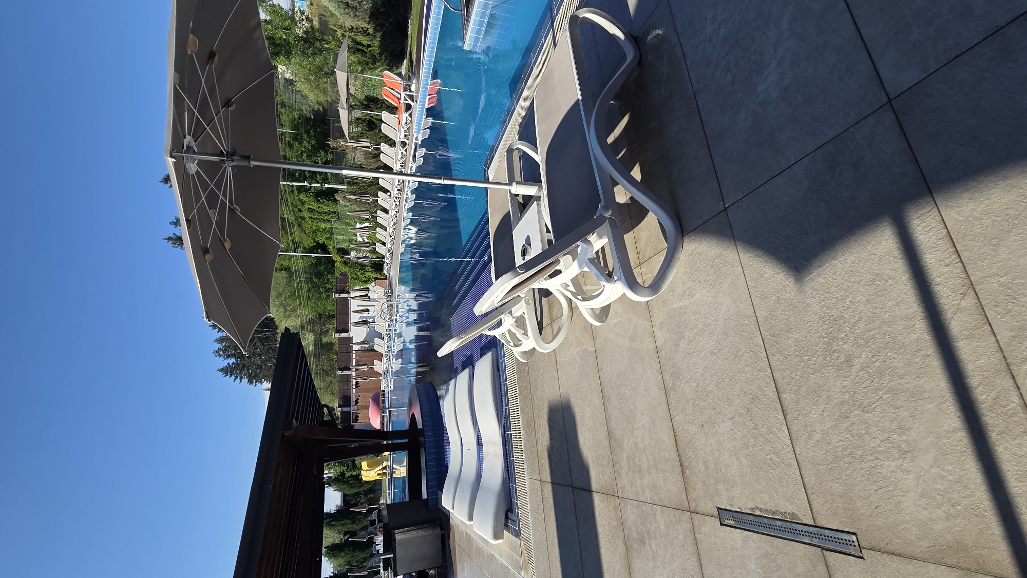 The image shows a resort pool area with clear blue water and lounge chairs on the tiled deck. In the foreground, two white and grey lounge chairs are positioned near a small table, with a large umbrella casting a shadow. Several other lounge chairs and umbrellas are visible along the edge of the pool and further back in the scene. The pool has tiled steps leading into it, and there are some inflatables visible in the shallow end. A covered seating area with a bar is on the left side of the image. The background shows trees and a clear blue sky. The resort appears to be located in Bania, Bulgaria.