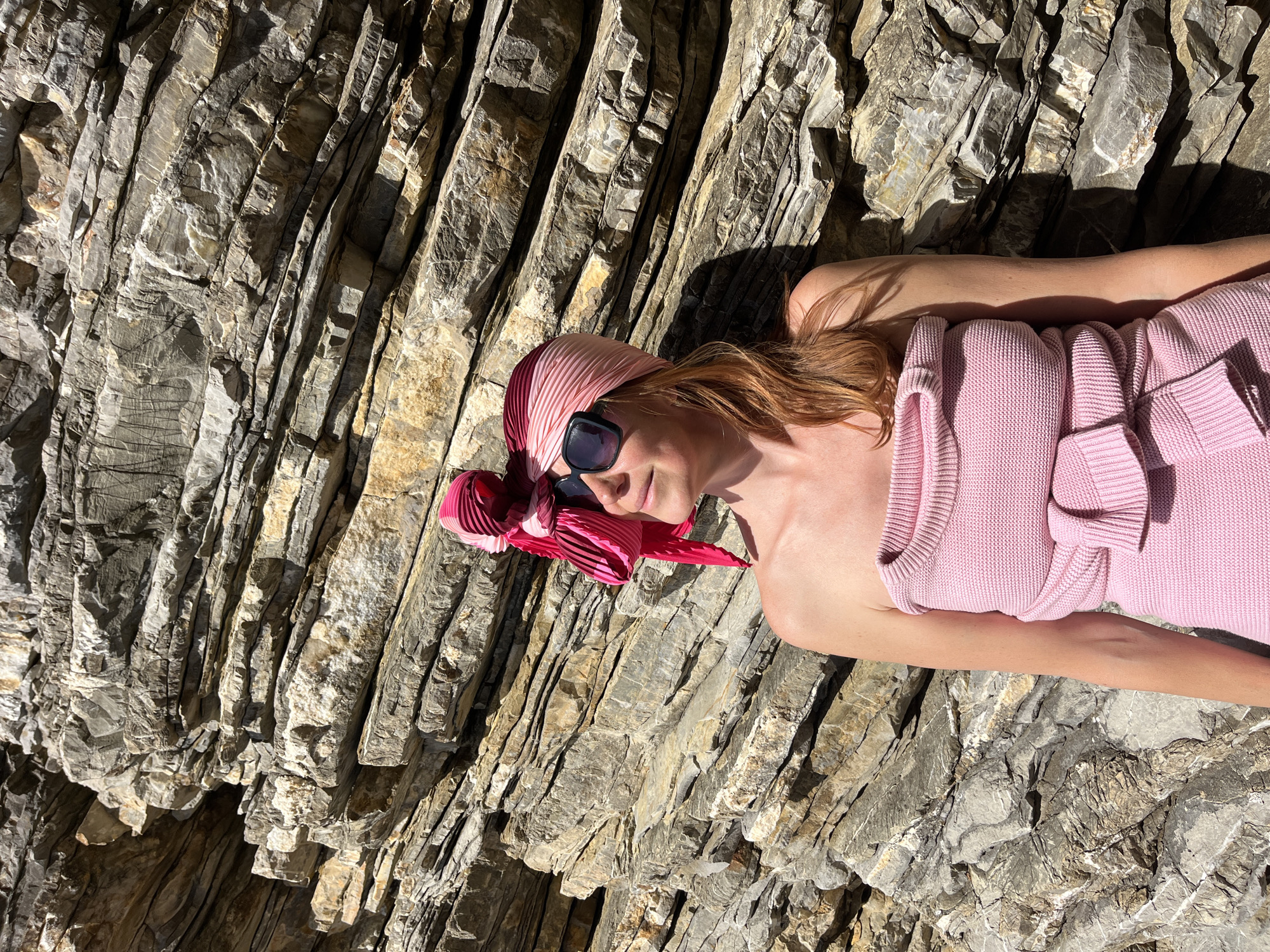 The image shows a woman lying on a rocky surface. The woman is wearing a pink headscarf and sunglasses. She is also wearing a pink dress. The rocky surface appears to be a rock formation. The location is likely a beach area in Budva, Montenegro, known for its rocky beaches and coastline.