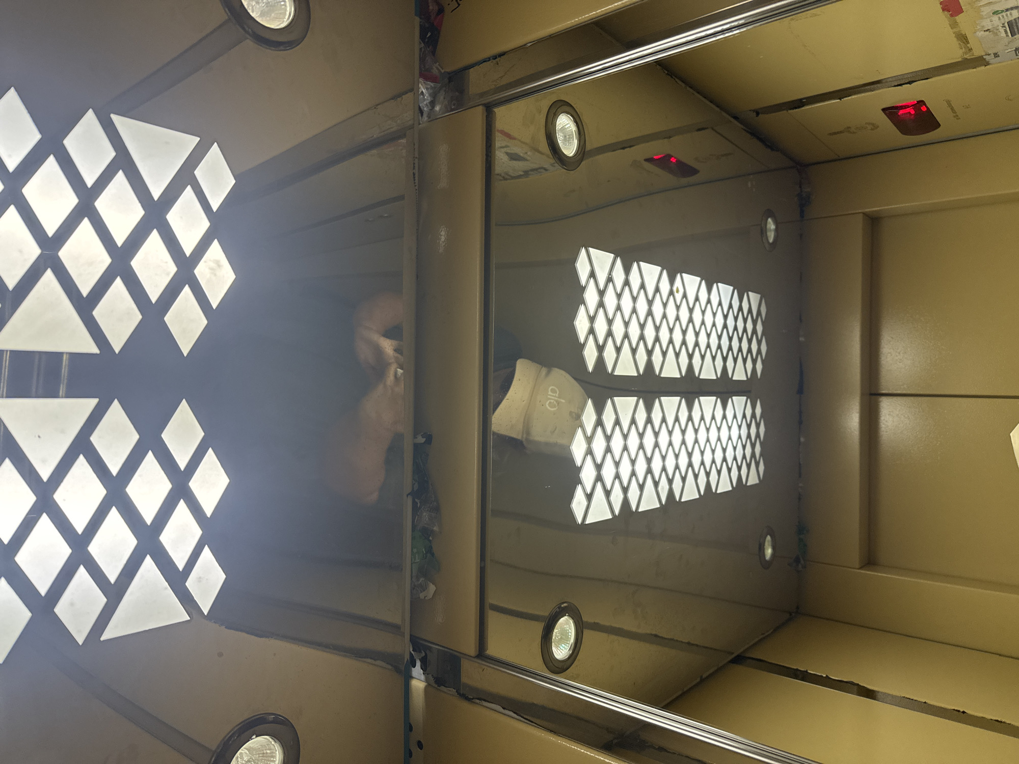 An overhead view looking up inside a yellow elevator car in Bishkek City, Kyrgyzstan. The ceiling features a diamond-patterned light fixture. A trash-strewn ledge is visible above the reflective mirrored walls. The mirrored walls reflect the interior of the elevator, including the ceiling lights and a red indicator light. The doors appear to be closed.