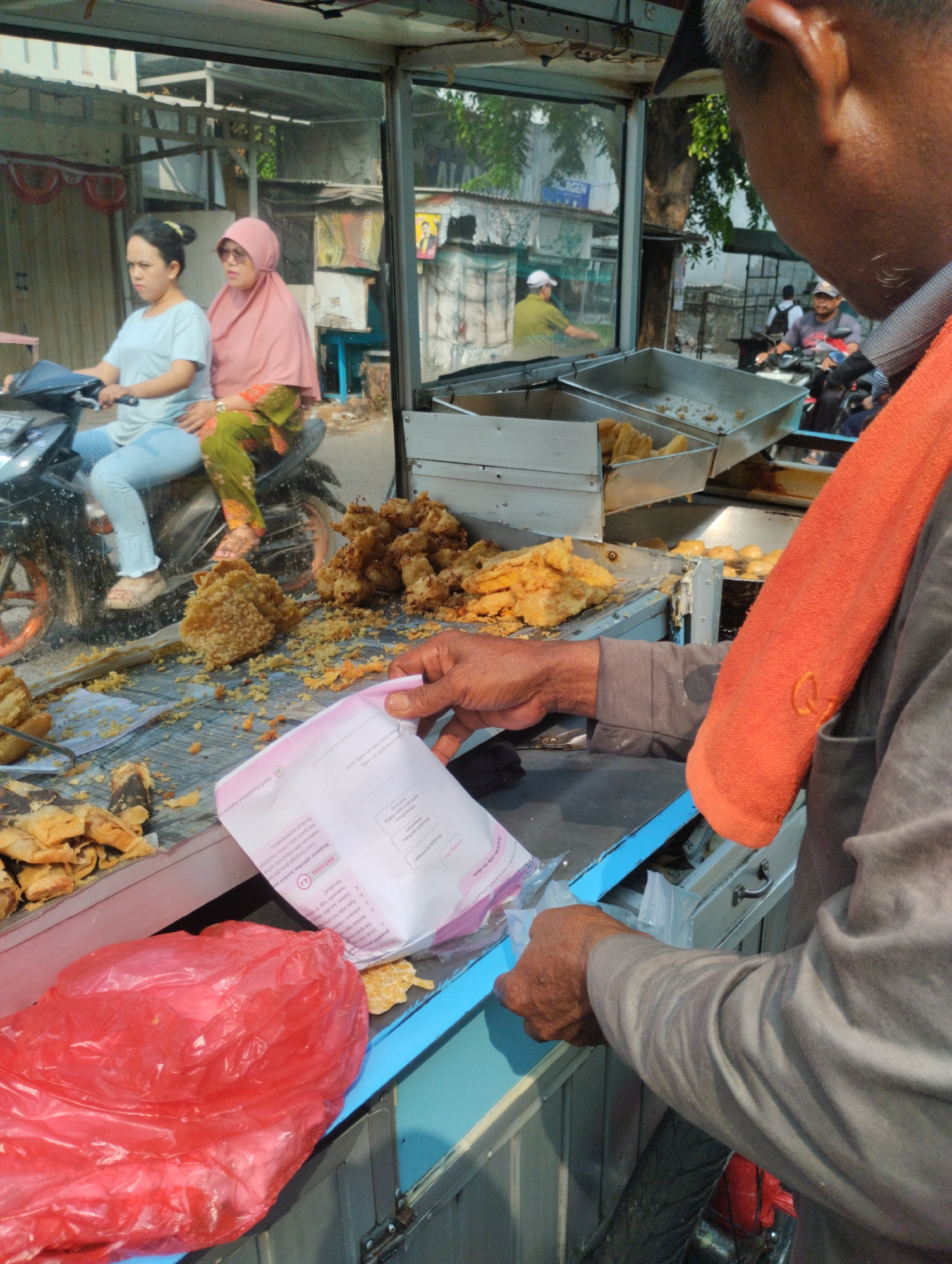 This is a medium shot, eye-level, outdoor photograph taken in Bekasi, Indonesia. The scene depicts a street food vendor, a man in the foreground wearing a grey shirt and an orange towel around his neck, interacting with a customer. The vendor is holding a pink and white paper with text on it, and appears to be in the process of serving food. Behind the vendor, a glass partition reveals a food stall with trays of fried items. Two women are visible on a motorcycle in the background, and other people and vehicles are present further down the street. The environment suggests a busy, urban setting with typical street infrastructure.