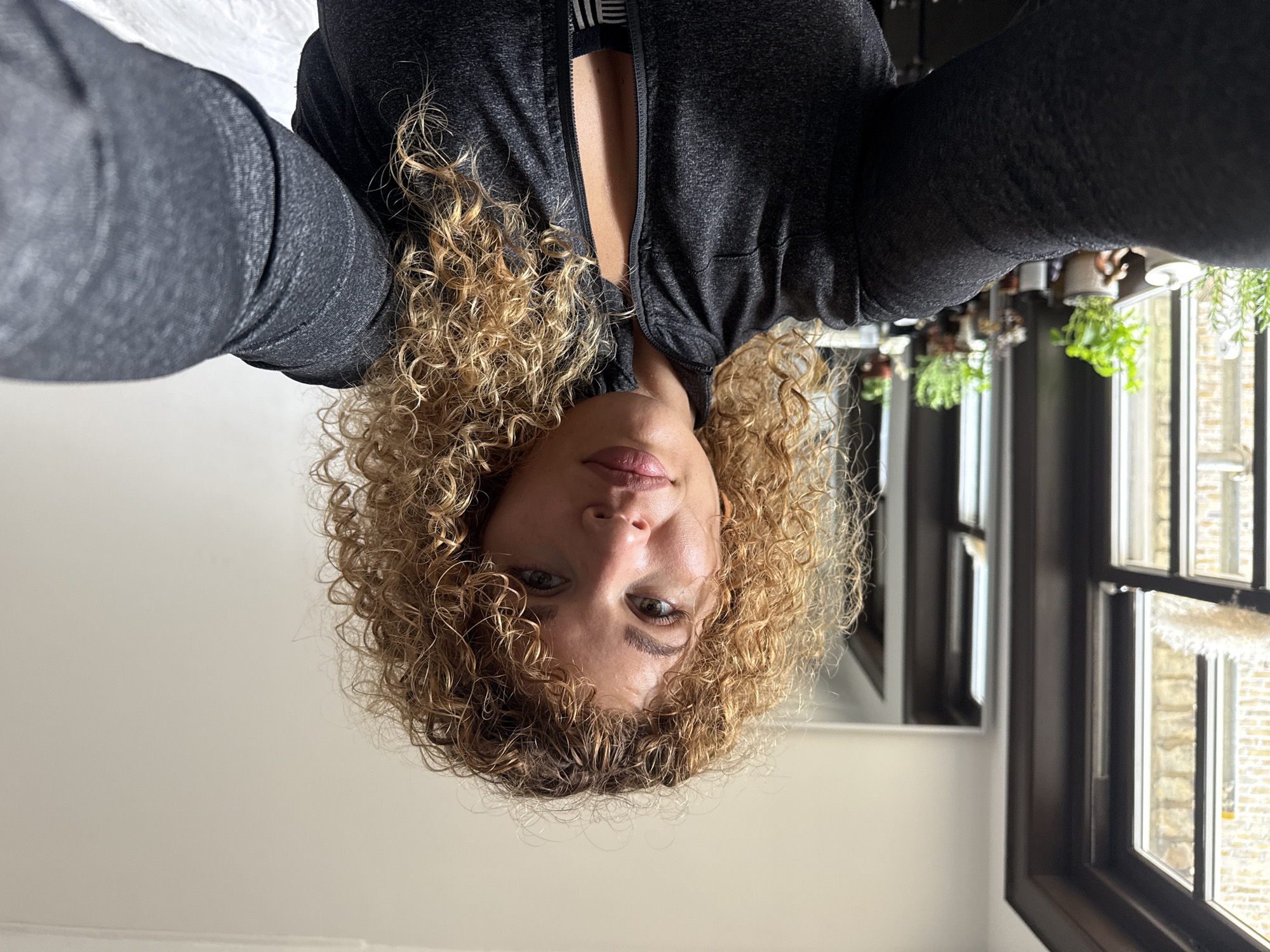 A woman with curly blonde hair takes a selfie in a room. She is wearing a dark grey zip-up jacket over a sports bra. The background shows a window with plants on the sill and a mirror reflecting more of the room. The scene appears to be indoors, likely a residential setting, given the personal items and decor. There is no indication that this location is in London, United Kingdom.