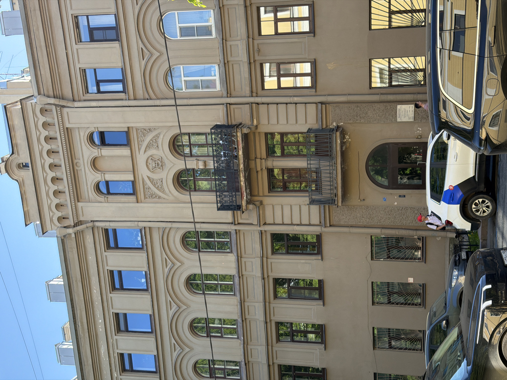 This image depicts a sunny street scene featuring a multi-story building with classical architecture, likely in Saint Petersburg, Russia. The building's facade is a light, cream-colored stucco, adorned with ornate moldings, cornices, and decorative carvings, including arched window frames and detailed motifs. It features a variety of windows: some rectangular with dark frames, and others with arched tops, some framed in white. Many windows reflect the bright blue sky and the foliage of unseen trees. Two wrought-iron balconies are visible on the building's middle floors.
Parked alongside the building are several cars. In the foreground on the right, a dark-colored car is partially visible. Adjacent to it is a white and blue vehicle, possibly a service car, with the number "57" and some blue text on its side. A person wearing a red cap, a light-colored top, and a backpack is seen standing near the white and blue car. Further down the street, other cars are also parked.
The lower windows on the right section of the building are protected by decorative metal grates. An arched entranceway is visible on this side of the building, above which a white rectangular sign with illegible text is affixed to the wall. The clear blue sky dominates the upper part of the image, and the strong sunlight casts shadows, indicating it is midday or early afternoon. Thin black wires extend across the upper portion of the frame.
