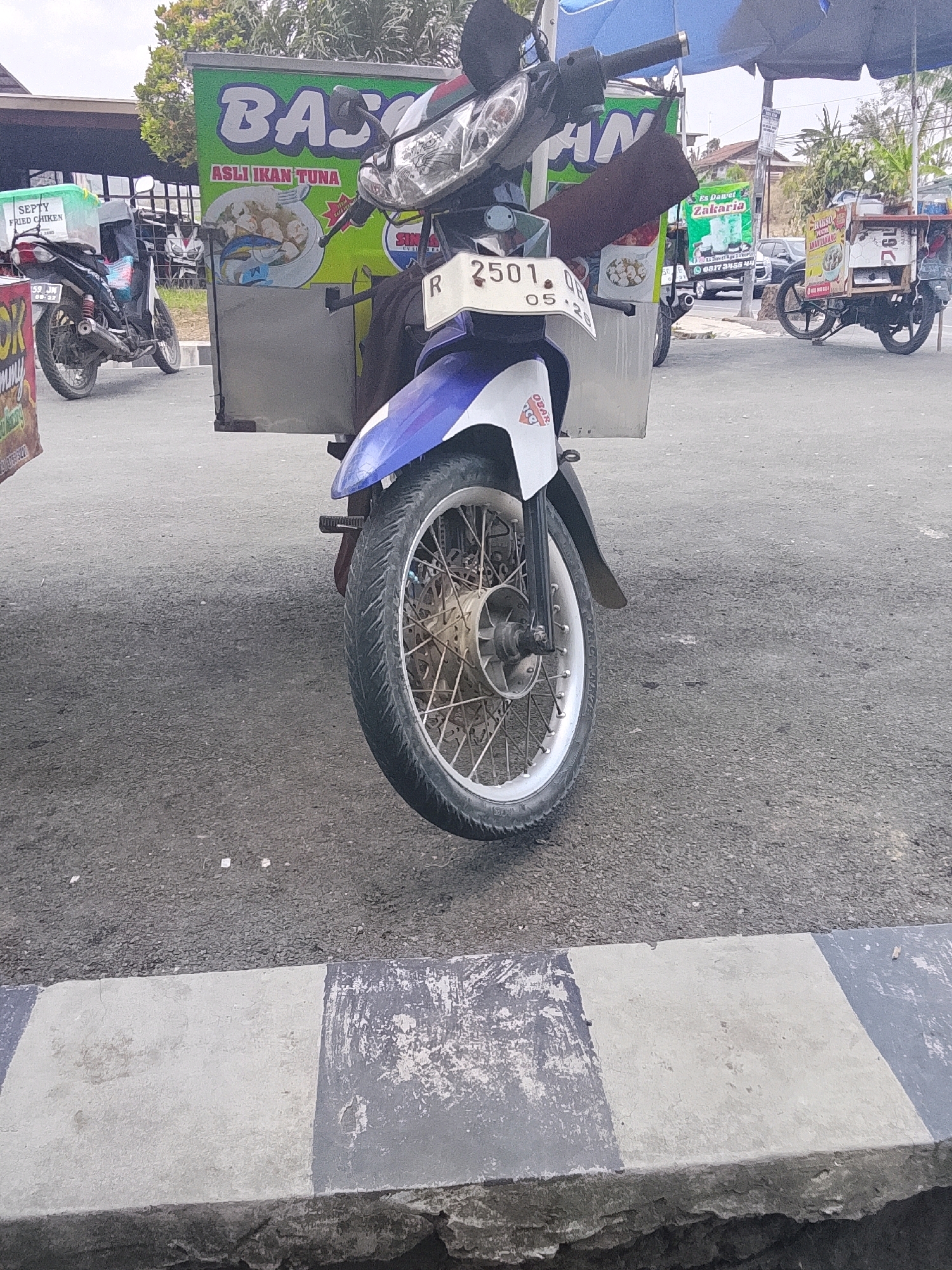 The image captures a street scene in Karangpucung, Indonesia, focusing on a motorcycle with a food cart attached. The motorcycle, predominantly white and blue, is parked on an asphalt road. Its front wheel and part of the handlebars are visible. A license plate reading "2501 OB 05-25" is attached to the front of the motorcycle. Behind the motorcycle, a green and white food cart is mounted, featuring text that appears to be advertising food items. To the left, another motorcycle is visible, also with a food container attached, labeled "SEPTY FRIED CHICKEN". Further in the background, buildings and other vehicles are present, suggesting a commercial or residential area. The overall environment appears to be outdoors during the daytime, with a paved road and a sidewalk in the foreground.