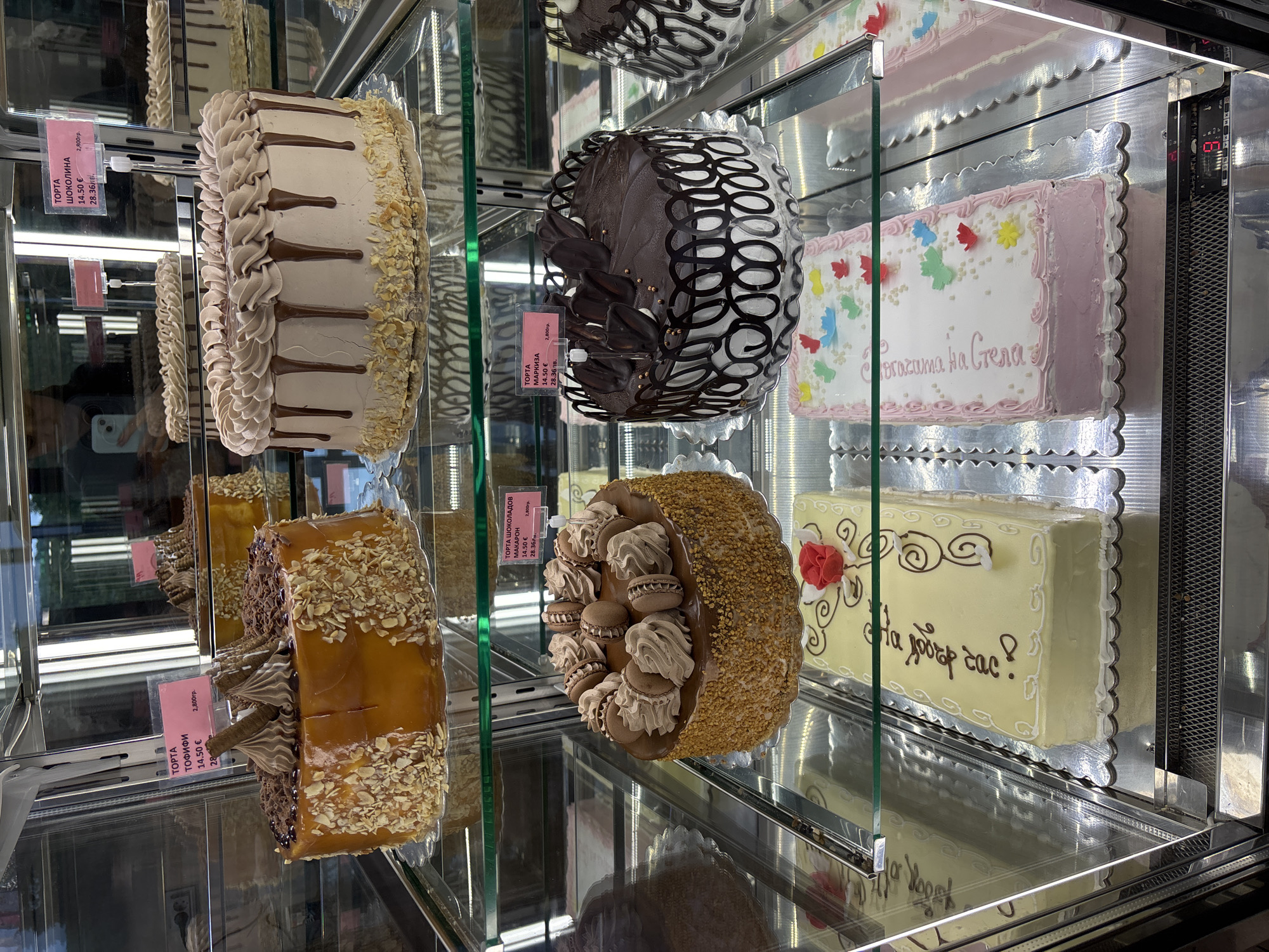 This image shows a refrigerated display case containing several cakes. The cakes vary in style and decoration. A price tag is visible on some cakes with information including "14.50 €", and "2,800гр.". The text on some price tags says "TOPTA ШОКОЛАДОВ" and "МАРКИЗА". The sign also shows a price in a currency besides Euros. The text on one of the cakes reads "Бгачата на Стела". These cakes are likely for sale in a bakery or confectionery store. Given the currency used in the price tag, this is potentially located in Gorna Malina, Bulgaria.