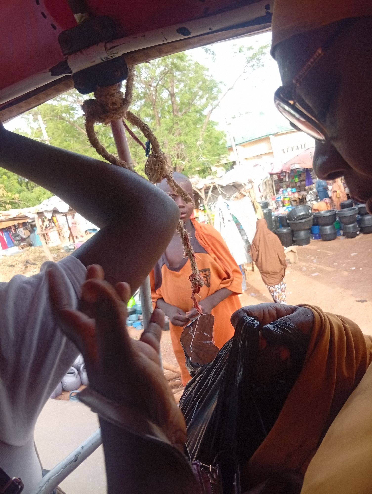 This image captures a candid scene from within an open-sided vehicle, looking out onto a bustling outdoor market in Dutse, Nigeria, during bright daylight hours. The vehicle's red ceiling structure, white metal bars, and a thick, knotted rope are prominent in the foreground, framing the view.
In the immediate foreground, fragments of several individuals are visible. To the left, a person's forearm, clothed in a white t-shirt sleeve, and their hand are partially in frame. To the right, the side profile of a person's head, wearing dark-framed glasses and a light-colored cap, is seen, looking towards the market. This person's arm and side, dressed in a brown-toned garment, clutch a black plastic bag.
The central subject is a young child with dark skin, wearing a bright orange shirt, who stands in the midground. The child looks directly at the viewer with a neutral expression, appearing to hold a small, string-like object in their hands. Behind the child, another individual in a brown head covering and patterned trousers is partially visible, walking away from the camera.
The background reveals a lively market setting with numerous makeshift stalls under the clear sky. Goods such as stacks of black plastic buckets and other household items are displayed for sale on dusty ground. Green trees dot the landscape beyond the market, and some light-colored buildings are visible in the distance. The overall scene depicts a typical vibrant street market environment. There is no visible text in the image.
