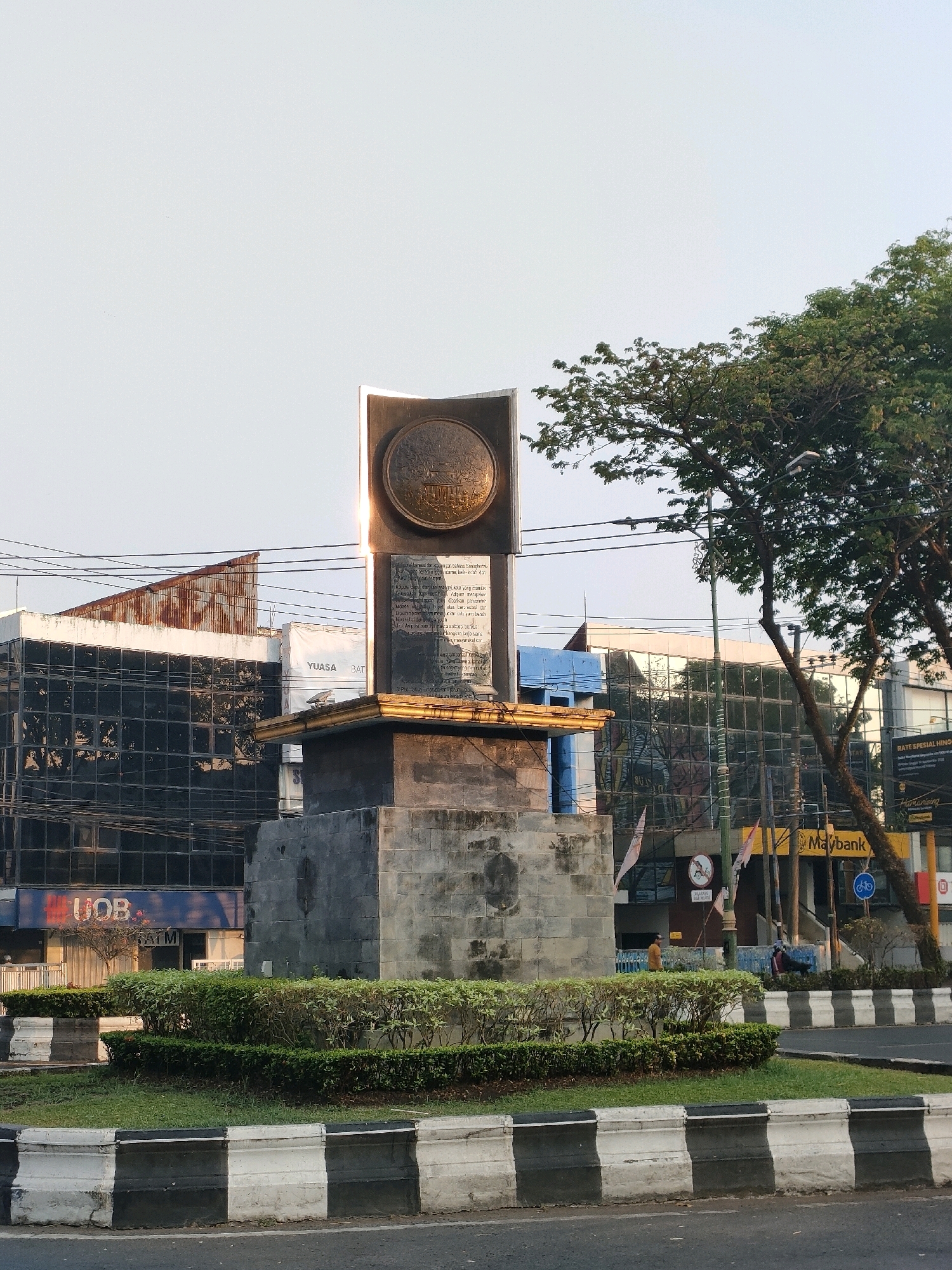 The image shows a monument in the city of Banjarmasin, Indonesia, at a street intersection. The monument is made of stone and metal, with an inscription and a circular emblem on its upper part. It is surrounded by a well-maintained garden. In the background, there are several modern buildings with glass facades, one of which has the "UOB" bank logo visible. A large tree with lush green foliage is to the right of the monument. The sky is clear and light, suggesting it is daytime. Traffic barriers with black and white stripes are visible along the road. Several flags are seen on poles. A few people are visible walking in the background, and a motorcycle is parked near the Maybank building. The overall scene depicts an urban environment with a prominent public monument.