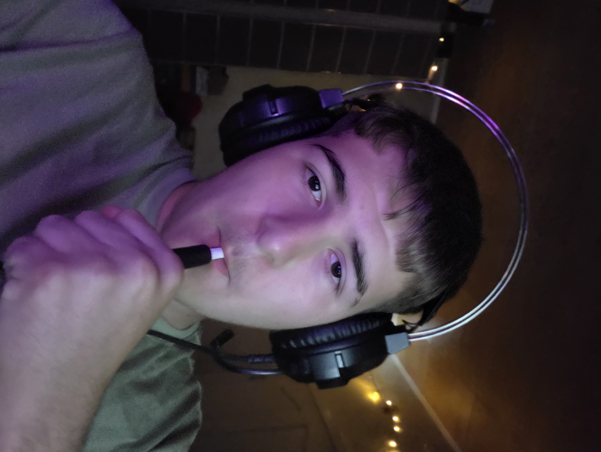 The image is a close-up, selfie-style photograph of a young Caucasian male wearing black headphones with a silver headband. He is wearing a green t-shirt and has a light-colored object, possibly a stylus or electronic cigarette, in his mouth. The lighting in the image is purple, suggesting artificial light sources, and there are small, out-of-focus lights in the background. The background appears to be indoors, with a dark wall and some shelving visible on the left. The overall impression is that of a casual, personal moment captured at home. There is no information to suggest the image is located in Aleksandrovo, Bulgaria.