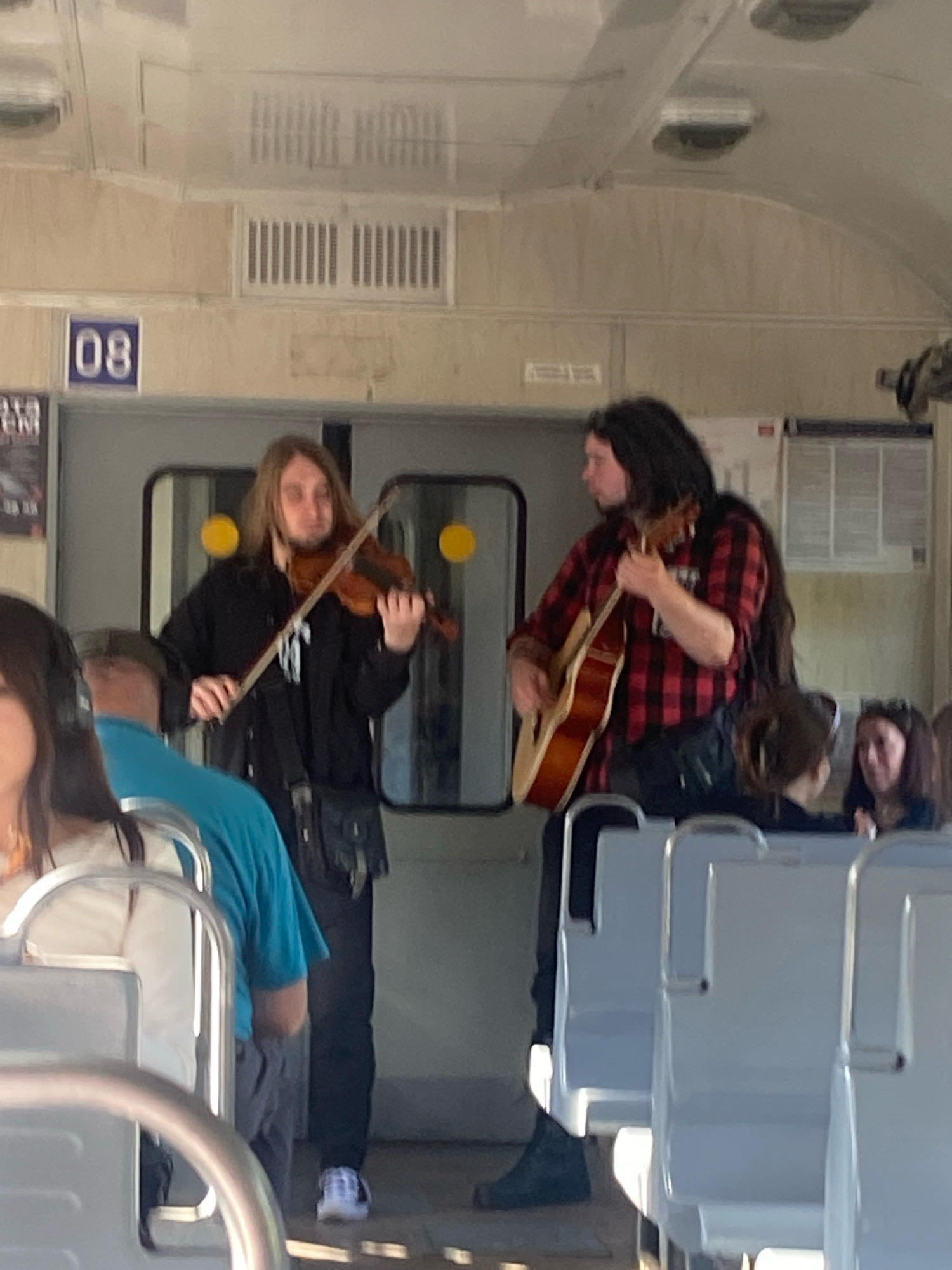 Two individuals are performing music inside a train car, possibly in Levashovo, Russia, judging by the context of the surrounding images. The person on the left, with long blonde hair, is playing a violin, holding the bow in their right hand and the violin under their chin. They are wearing a black hoodie and dark pants, with a dark pouch or bag hanging from their waist. The person on the right, with long dark hair, is playing an acoustic guitar. They are wearing a red and black plaid shirt and dark pants, with a dark bag slung across their chest. The train car interior is visible, with rows of light-colored seats. In the background, there are windows and doors, and a sign with the number "08" is mounted on the wall. Other passengers are partially visible in the foreground and background, some seated and some standing. The lighting suggests it is daytime.