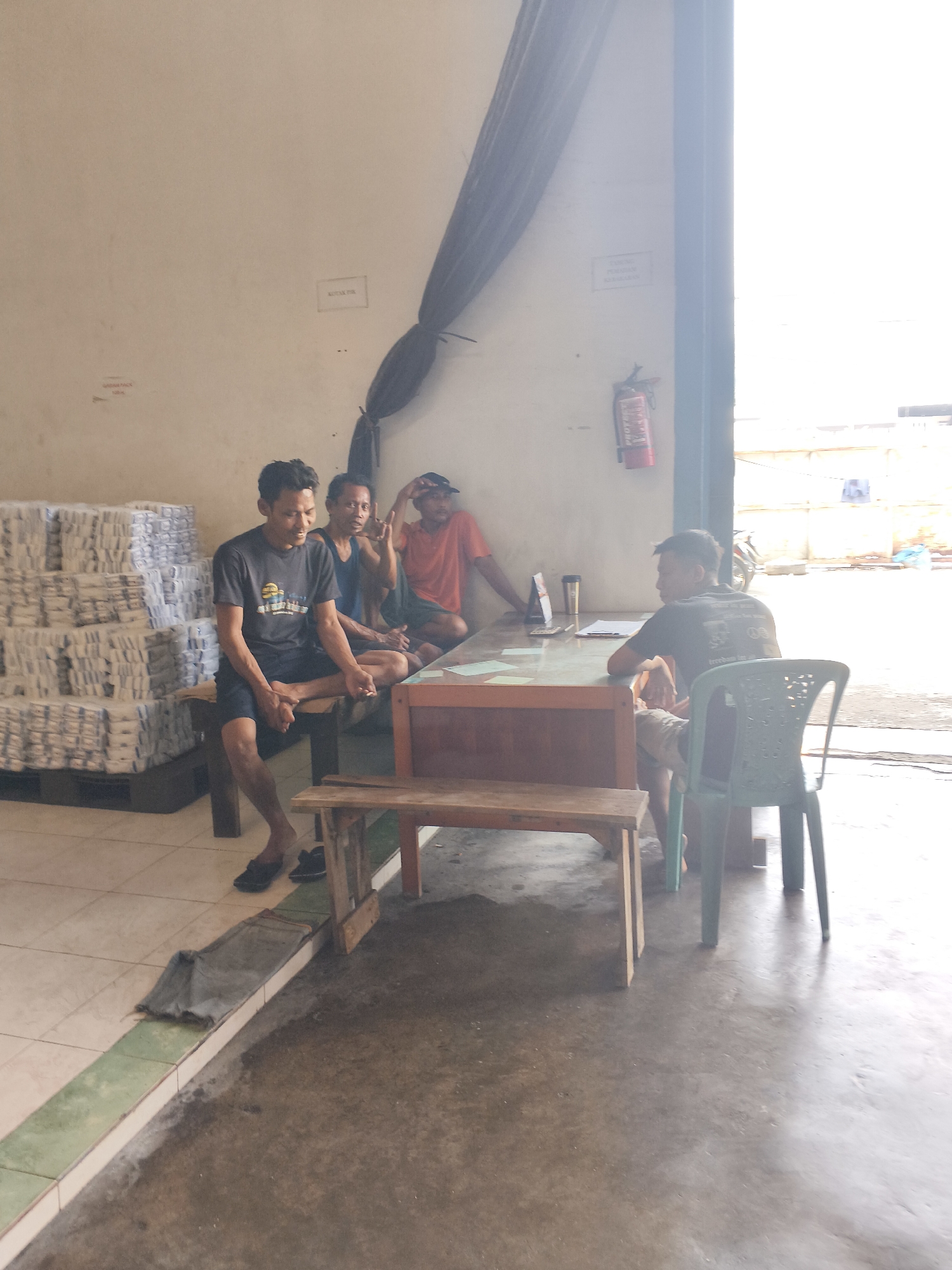 Four men are gathered in an indoor industrial or warehouse setting. Three men are seated at a table, and one man is seated on a bench. A stack of white packages is visible to the left of the frame. A red fire extinguisher is mounted on the wall to the right. The lighting suggests it is daytime, with bright light coming from an open area to the right, possibly an entrance or exit. The flooring appears to be concrete and tile. The men are casually dressed. The overall scene depicts a break or informal meeting.