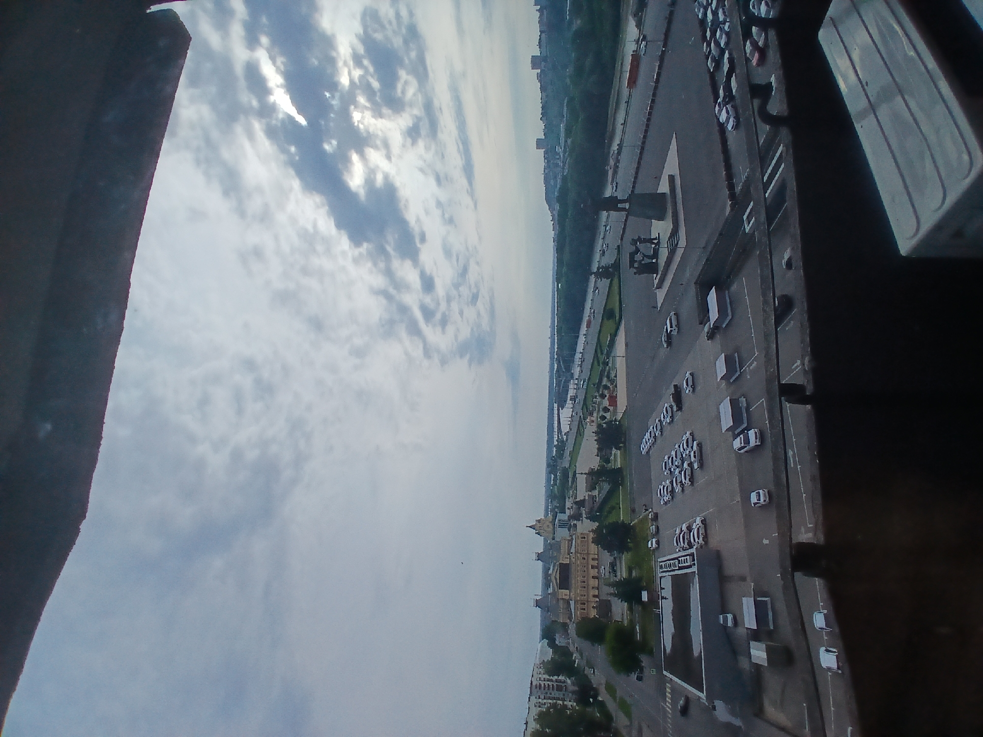 An aerial view of the city of Nizhny Novgorod, Russia, taken from an elevated vantage point. The scene captures a cityscape with a river in the background, a large plaza with a monument, a parking lot filled with cars, and various buildings. The sky is overcast with scattered clouds. In the foreground, there is a dark, blurred object which appears to be a part of a structure, possibly a balcony or window frame, obscuring the lower portion of the image.
The plaza features a prominent bronze monument depicting figures standing on a pedestal. Adjacent to the plaza is a wide road with moving traffic, bordered by a green expanse of trees. Across the road, there is a large, yellow building with a distinctive dome, likely a significant architectural landmark. Further in the distance, the river stretches across the horizon, with hints of buildings and greenery on the opposite bank.
A large parking lot occupies a significant portion of the mid-ground, filled with numerous white and silver cars parked in rows. Some of these cars are also visible in the lower foreground, parked in designated spots. Several small, grey utility structures or sheds are scattered within the parking lot. The overall impression is a busy urban environment under a cloudy sky.
