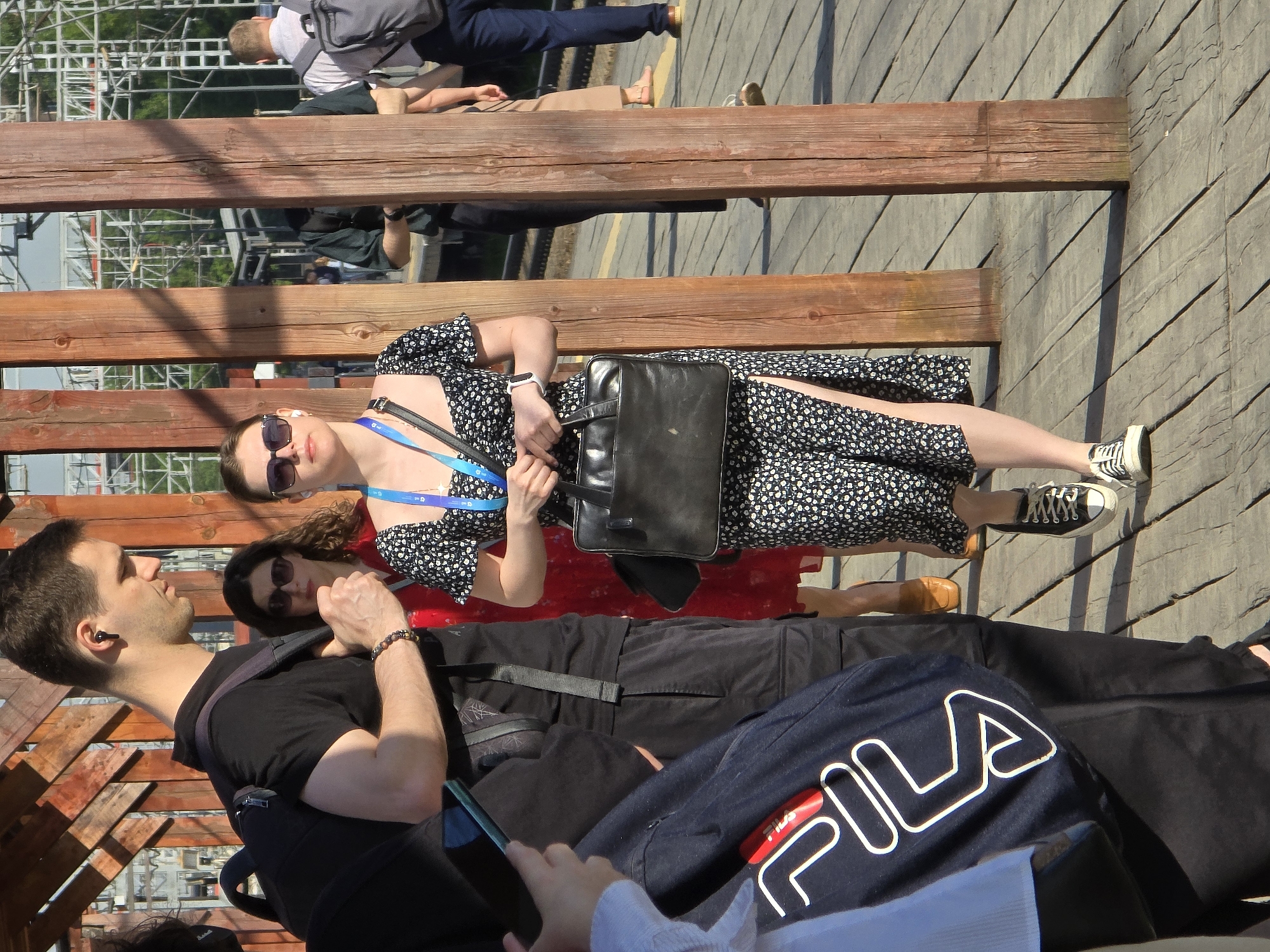 The image shows a group of people on a wooden bench. A woman wearing a floral dress, sunglasses, and sneakers is holding a black bag. Next to her, a man in a black shirt and backpack is looking towards her. Another woman wearing sunglasses and a red top is visible behind them. A blue Fila jacket is visible near the bottom of the image. The location is Khimki, Russia. Overhead, there's a metallic structure and pedestrian walkway. The surface below is paved.