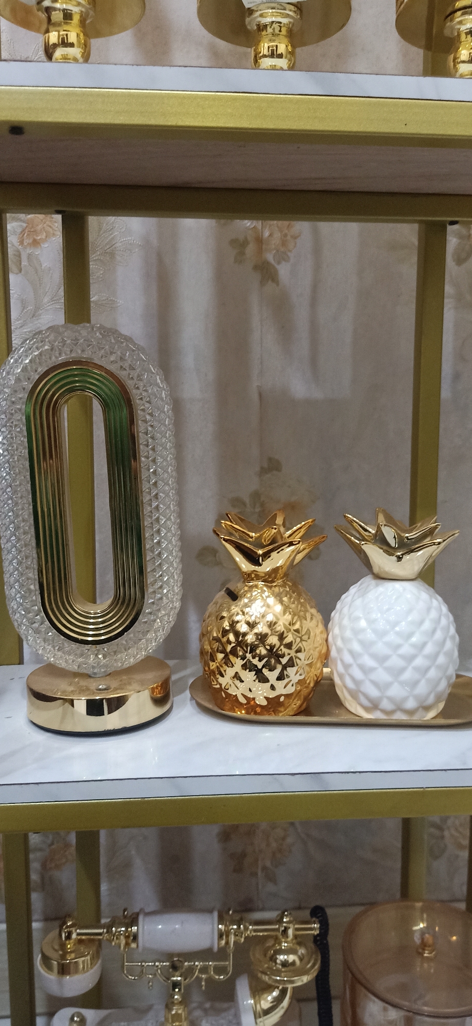 This image shows decorative objects on a shelf. On the left is a clear, oval-shaped object with gold-colored ribbed detailing inside, mounted on a gold base. To the right of this are two pineapple-shaped piggy banks on a gold tray. One piggy bank is shiny gold, and the other is white with a textured surface. In the background, a patterned curtain is visible. Below the shelf, a decorative gold and white rotary phone is partially visible, along with a gold cylindrical container. The scene appears to be indoors, possibly in a home or store, and is not identifiable as being in Lhokseumawe, Indonesia.