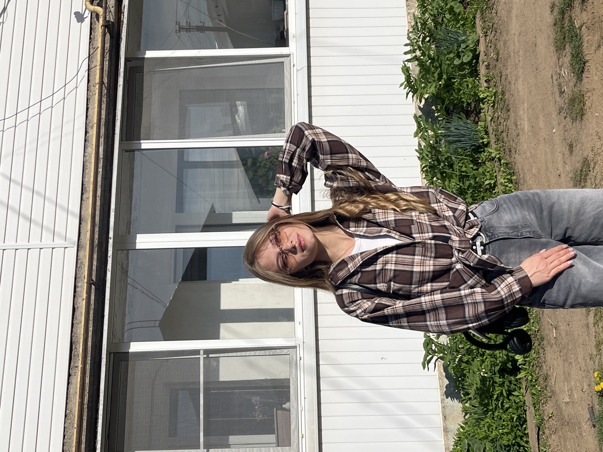 A young woman stands outdoors, posing for a photograph. She is wearing a brown and beige plaid shirt, a white t-shirt underneath, and gray jeans. She also has glasses on her face and a bracelet on her wrist. A pair of black headphones are laying on the ground near her.
The location appears to be the exterior of a building, possibly a house, with white horizontal siding and several windows. There is green vegetation growing along the side of the building. The ground is dirt and has some sparse patches of grass.
Based on the provided location, the setting is in Фоки, Russia, although there are no objects to verify this information.