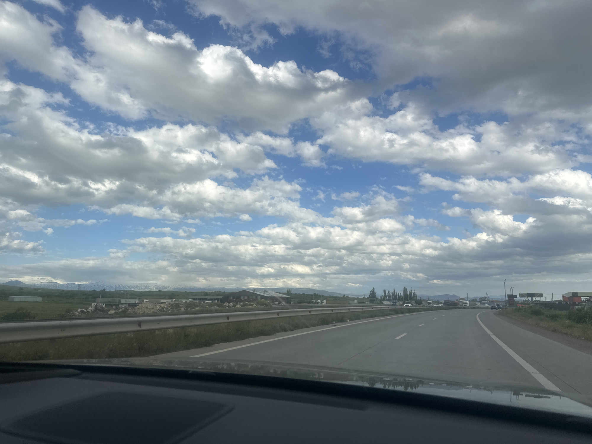 This photograph captures a daytime view from inside a vehicle, looking out onto a multi-lane highway that stretches into the distance. The dark dashboard and a reflection are visible in the immediate foreground.
To the left of the road, a metal guardrail runs alongside a grassy embankment. Beyond this, the landscape features flat, green fields dotted with various structures, including low-rise buildings, some appearing to be industrial, agricultural, or possibly under construction. In the far background, impressive snow-capped mountains rise beneath the horizon.
The highway itself curves gently to the right, with clear white lane markings visible. On the right side of the road, there are clusters of trees, what appear to be commercial or industrial buildings, and several parked or moving vehicles, including trucks. A distant sign on the right side of the road has barely legible text, possibly including "GAS".
The sky is a prominent element of the scene, filled with large, dramatic white and grey cumulus clouds interspersed with patches of clear blue sky, suggesting a dynamic weather pattern. The overall impression is one of travel through a diverse landscape, combining natural features like mountains and greenery with human infrastructure. The presence of snow-capped peaks aligns with the given location context of Proshyan, Armenia.
