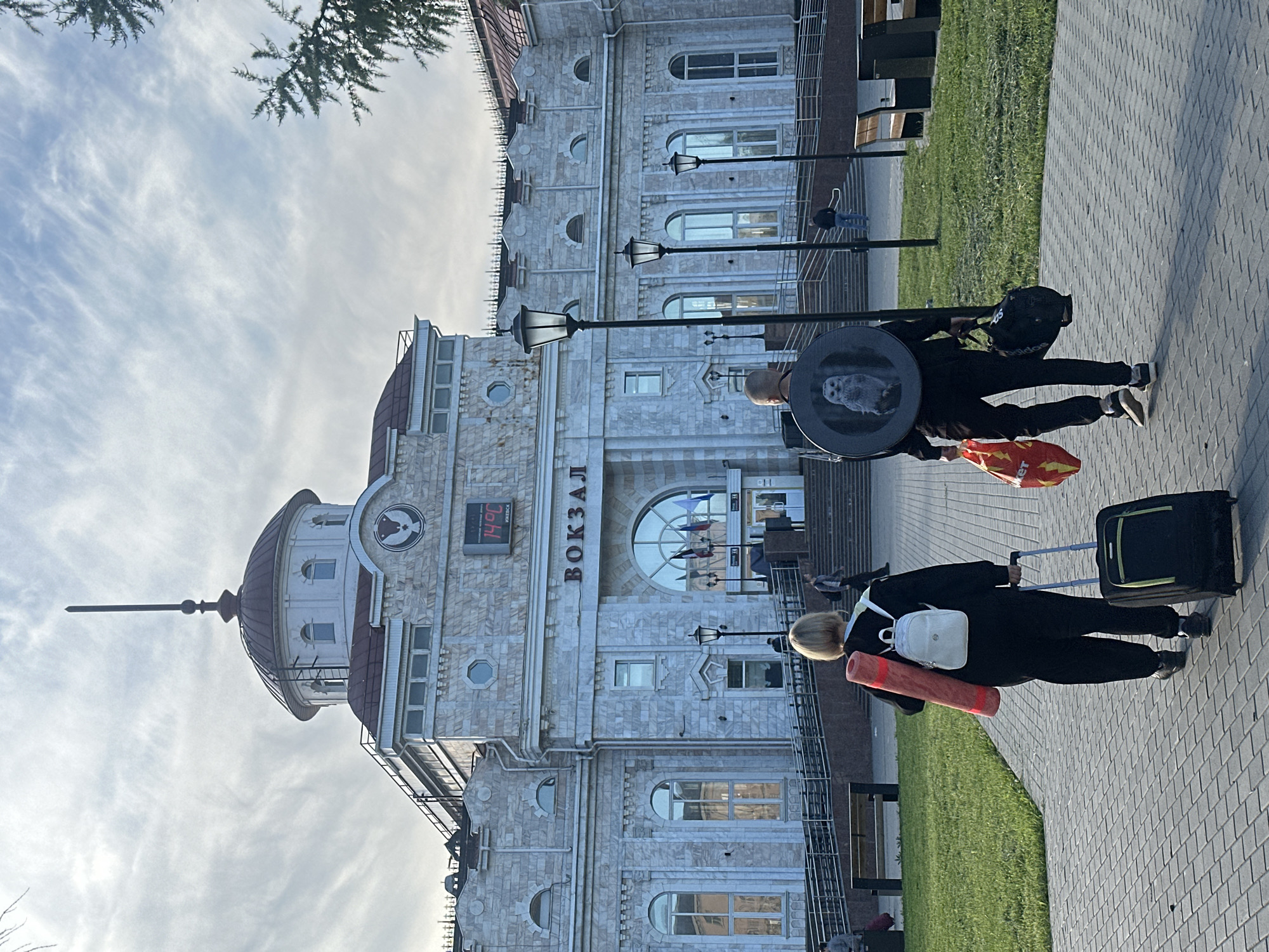 The image depicts an outdoor scene in front of a grand, light-colored stone building, identified as a "ВОКЗАЛ" (Vokzal, or station) in Izhevsk, Russia. The sky above is partially cloudy with wispy white formations.
Two individuals, viewed from behind, are walking along a paved pedestrian path away from the camera. The person closer to the viewer has blond hair, wears a dark outfit, a white backpack, and carries a rolled-up red object (possibly a mat) under one arm. She is pulling a black rolling suitcase with a neon green accent. The second person, positioned further down the path, also wears a dark outfit and carries a black backpack featuring an "Adidas" logo. She holds an orange shopping bag and a distinctive large circular object with a detailed white owl design on its surface, reminiscent of Hedwig from Harry Potter.
The station building is elaborate, constructed of light-colored, patterned stone blocks. It features numerous arched and rectangular windows. On the left side, a prominent dome with a tall spire extends towards the sky. A digital display on the building's facade clearly shows "14°C" and the city name "ИЖЕВСК" (Izhevsk). Several black, ornate lampposts line the paved path in front of the building, which is bordered by patches of green grass. A few tree branches are visible at the top of the frame. The scene appears to be set during the daytime, likely morning or late afternoon, as people move with their belongings.
