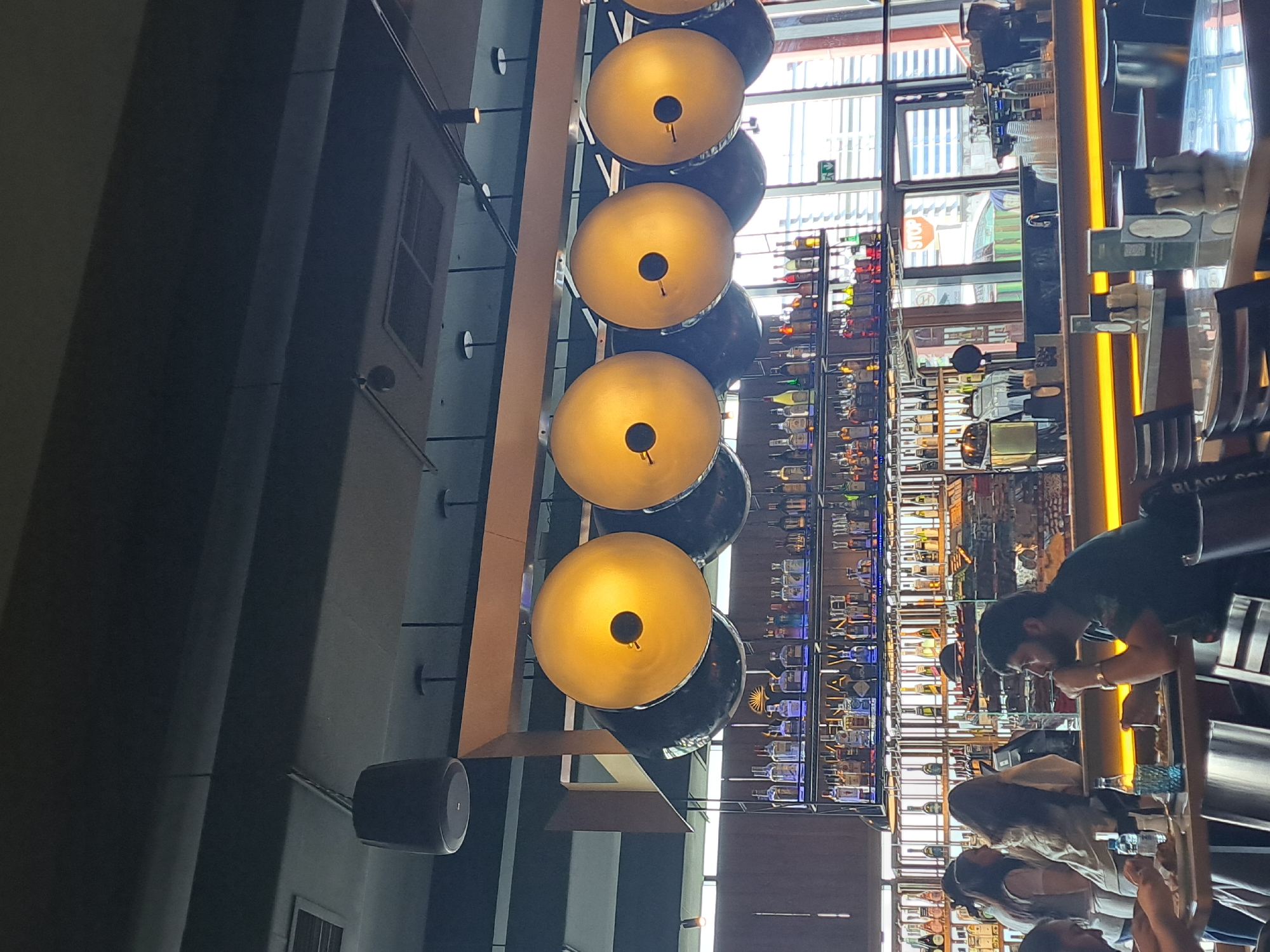 The image depicts the interior of a bar, likely in Yerevan, Armenia. The scene is composed of several key elements:
**People:**
* Patrons are seated at the bar, with a few people in focus. One man is wearing a green shirt. There are women with long hair. The people are interacting and enjoying themselves.
**Objects:**
* Light fixtures with round, yellow, globe-like lamps are installed, suspended in a vertical line.
* Bottles of various liquors are stocked on shelves behind the bar.
* Bar stools line the counter.
* Speakers are mounted on the ceiling.
**Actions:**
* People are sitting and engaging in conversation.
**Environment:**
* The setting is a modern bar.
* The bar has a view of what appears to be an urban street with a window filled with natural light.
* A shelving unit with bottles behind the bar is a prominent feature.