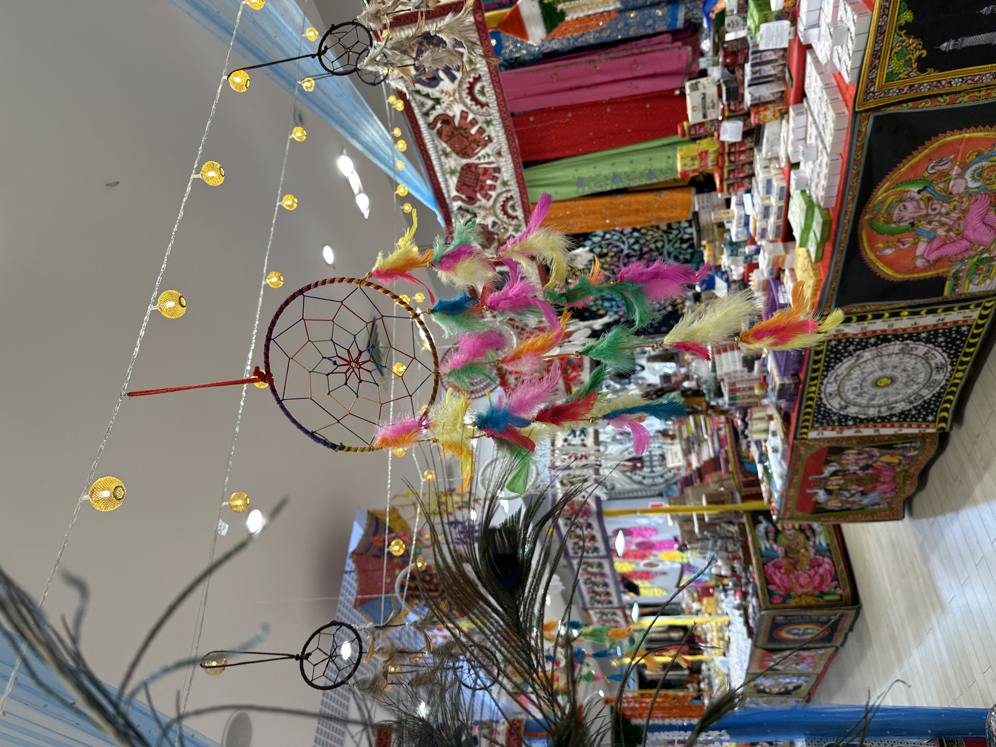 This vibrant image captures an array of cultural decorative items within what appears to be an indoor market or shop in Yekaterinburg, Russia. The scene is characterized by rich colors and intricate designs, creating a festive atmosphere.
In the foreground, a large, ornate dreamcatcher hangs prominently, adorned with a multitude of brightly colored feathers in shades of pink, yellow, green, blue, and orange. Its intricate webbing is visible within the hoop. Several smaller, simpler dreamcatchers, some black, are also scattered throughout the upper portion of the frame. Below and slightly to the right, a cluster of dark, iridescent peacock feathers adds another decorative element.
The background reveals a bustling display of goods. Colorful textiles, possibly saris or tapestries, are draped and stacked, featuring patterns like red, green, orange, and blue fabrics. One notable textile shows an ornate pattern with elephants. Numerous small, white and colored boxes are neatly stacked on shelves or tables, suggesting packaged products such as incense or spices. Further to the right, several artworks or tapestry-like hangings depict Hindu deities, including a prominent image of Ganesha and another figure sitting on a lotus flower, likely Lakshmi. A circular mandala-like design is also visible on a dark background.
The setting is illuminated by strings of warm yellow orb lights hanging from the ceiling, contributing to the inviting ambiance. No people are visible, but the arrangement of goods suggests an active retail environment, implying shopping or browsing as the primary activity. The strong cultural motifs, particularly the Hindu deities and specific textiles, indicate a specialty shop focusing on Indian or South Asian imported goods. No discernible text is clearly readable on any of the products or displays. The image's vertical orientation suggests it was captured and later rotated.
