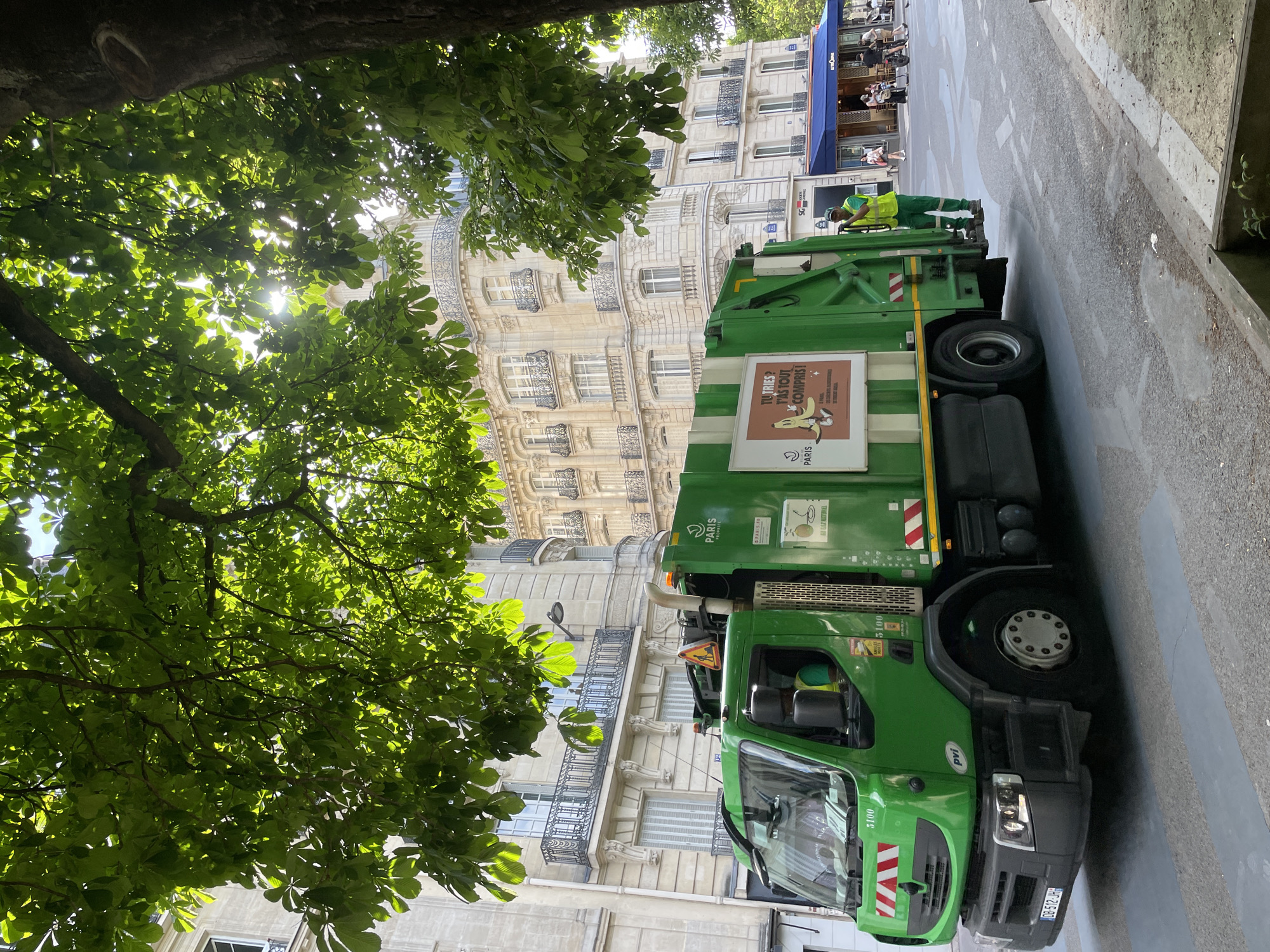 An urban street scene in Paris, France, captured during what appears to be a bright, sunny daytime. The image is oriented vertically but depicts a horizontally laid-out street. The primary subject is a green garbage truck belonging to the city of Paris, positioned on the right side of the frame. A garbage collector, wearing a green uniform and a high-visibility yellow-green vest, stands at the rear of the truck, possibly engaged in collection duties or taking a break.
The truck features various markings and text. On its side, a large brown and white sign displays a cartoon character resembling a dog (perhaps Gromit-like) with a banana peel, accompanied by the French text "T'U TRIES? T'AS TOUT COMPRIS!" (You sort? You've understood everything!). Other visible text on the truck includes "PARIS PROPRETE", "GAZ NATUREL", "PARIS", "5100", "pvi", and the license plate "OB-512-JH".
In the background, behind the truck, stand multi-story buildings characteristic of Parisian Haussmannian architecture, adorned with ornate balconies and intricate facade details. Sunlight illuminates these buildings. Further down the street, on the left, an outdoor cafe with blue awnings is visible, where several people are seated, suggesting an active street environment.
The upper portion of the frame is dominated by dense green foliage from overhead trees, through which bright sunbeams filter, creating dappled light and a sense of natural canopy over the street. The street pavement shows some markings and a general urban appearance. The combination of the architecture, the specific text on the truck, and the cafe ambiance clearly places the scene in Paris.
