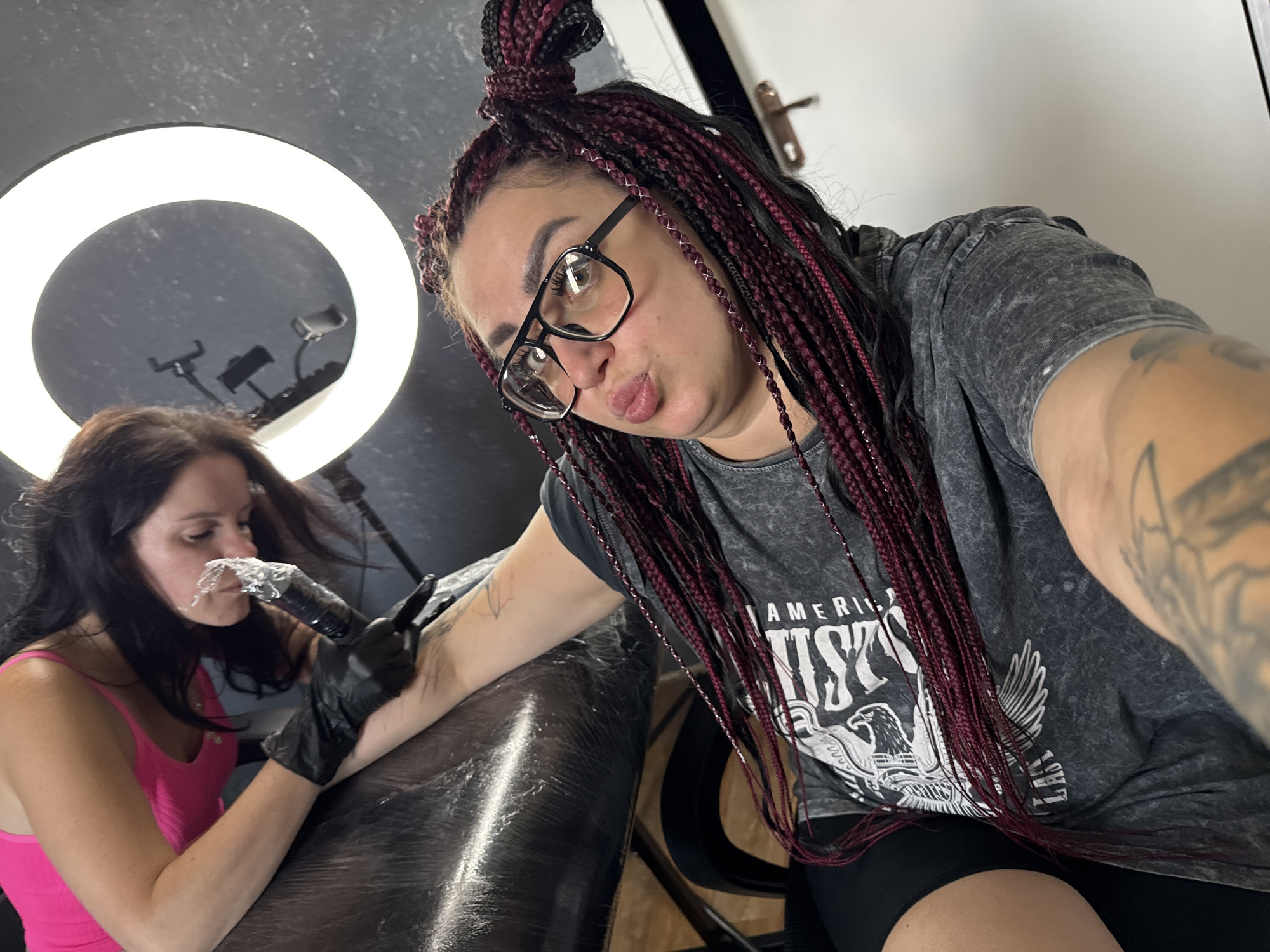 In Tolyatti, Russia, a woman with braided burgundy hair and black-rimmed glasses is taking a selfie. She is wearing a grey t-shirt with a graphic print and black shorts. Her mouth is puckered as if blowing a kiss. In the background, another woman, wearing a bright pink tank top and black gloves, is tattooing her arm with a tattoo gun. A ring light is visible in the upper right corner of the image. The scene appears to be a tattoo parlor.