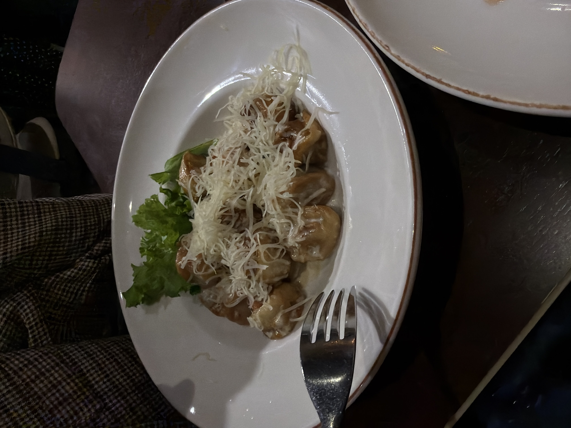 A close-up, eye-level shot shows a white plate filled with food, likely dumplings or gnocchi, covered in shredded cheese and a light sauce, served with a small garnish of green lettuce. A metal fork is positioned to the left of the plate, with its tines partially inserted into the food. The plate is set on a dark, wooden table, and a person's legs, dressed in a brown and beige checkered pattern, are visible in the upper left corner of the frame. The lighting is dim, suggesting an indoor setting, possibly a restaurant.