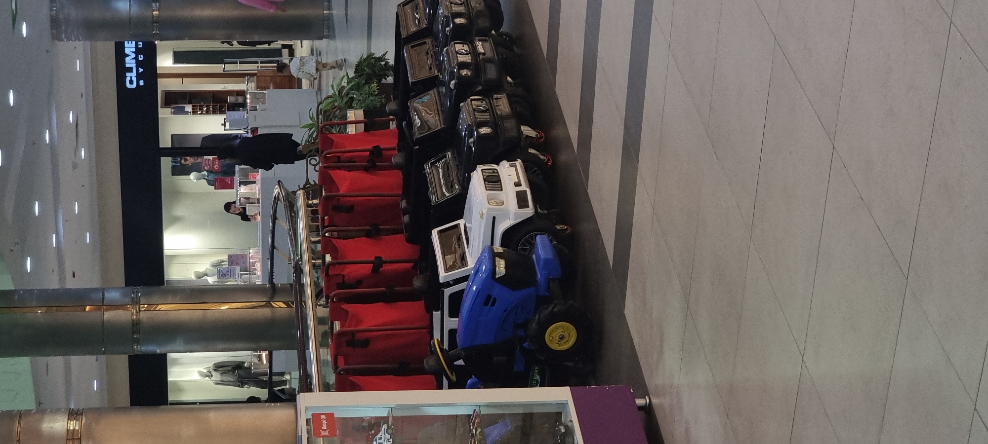 The image shows a row of toy cars, likely for rent, lined up in what appears to be a shopping mall in Aktobe, Kazakhstan. In the foreground, a blue toy tractor is parked next to a white toy SUV. Behind these, a row of black toy SUVs are visible. A metal railing with red fabric attached runs above the toy cars. In the background, mannequins are displayed in store windows, and a person is seen standing at a counter. The floor is tiled. The overall scene suggests a recreational area within a commercial establishment.
