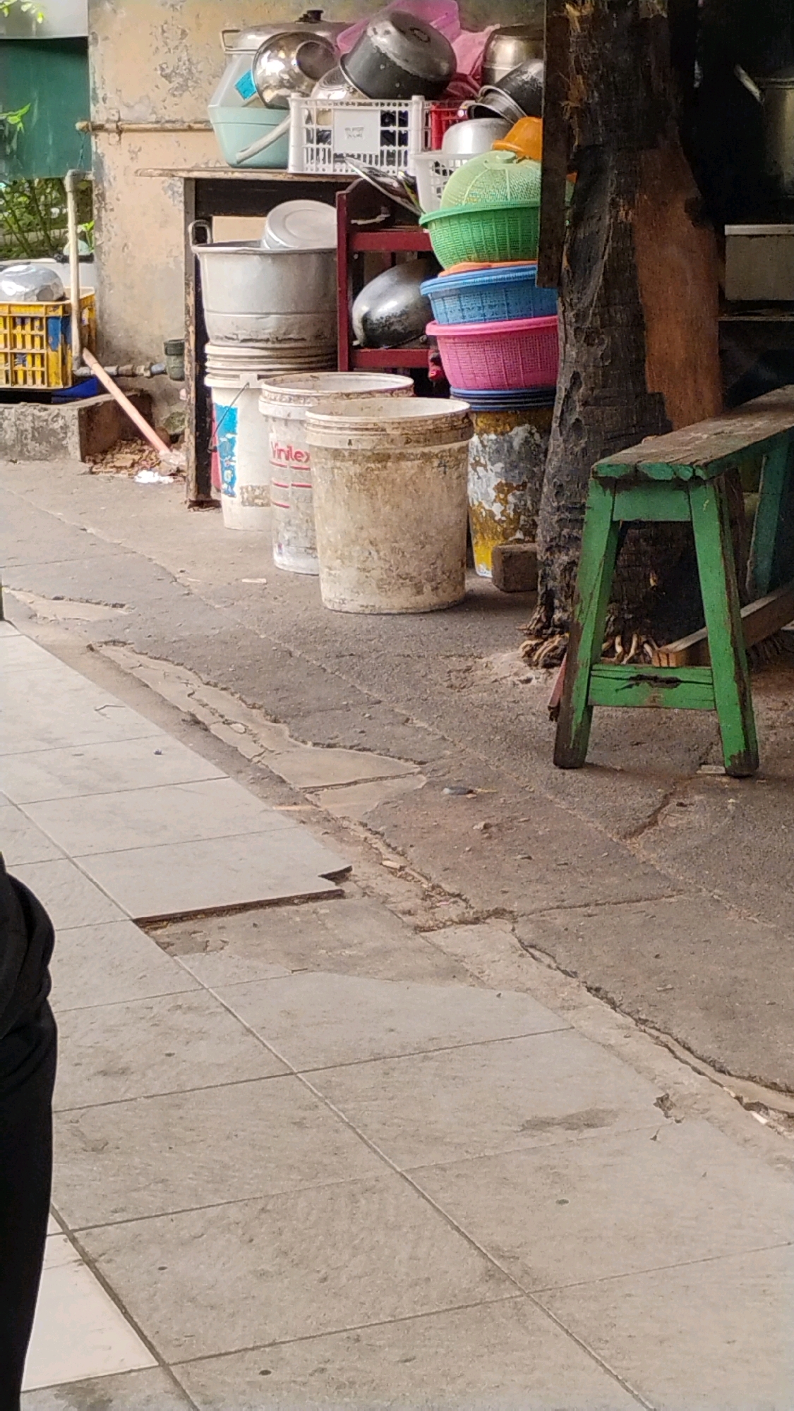 The image shows an outdoor scene with several stacked plastic baskets and buckets in front of a building. The building has a distressed, weathered wall. In the foreground, a tiled walkway leads away from the viewer. To the right, a rustic green wooden stool is visible. The ground is a mix of asphalt and cracked concrete. The overall impression is one of a busy, possibly commercial, area with storage of goods. The scene suggests an urban environment, likely in Central Jakarta, Indonesia, given the typical streetscapes and materials seen.