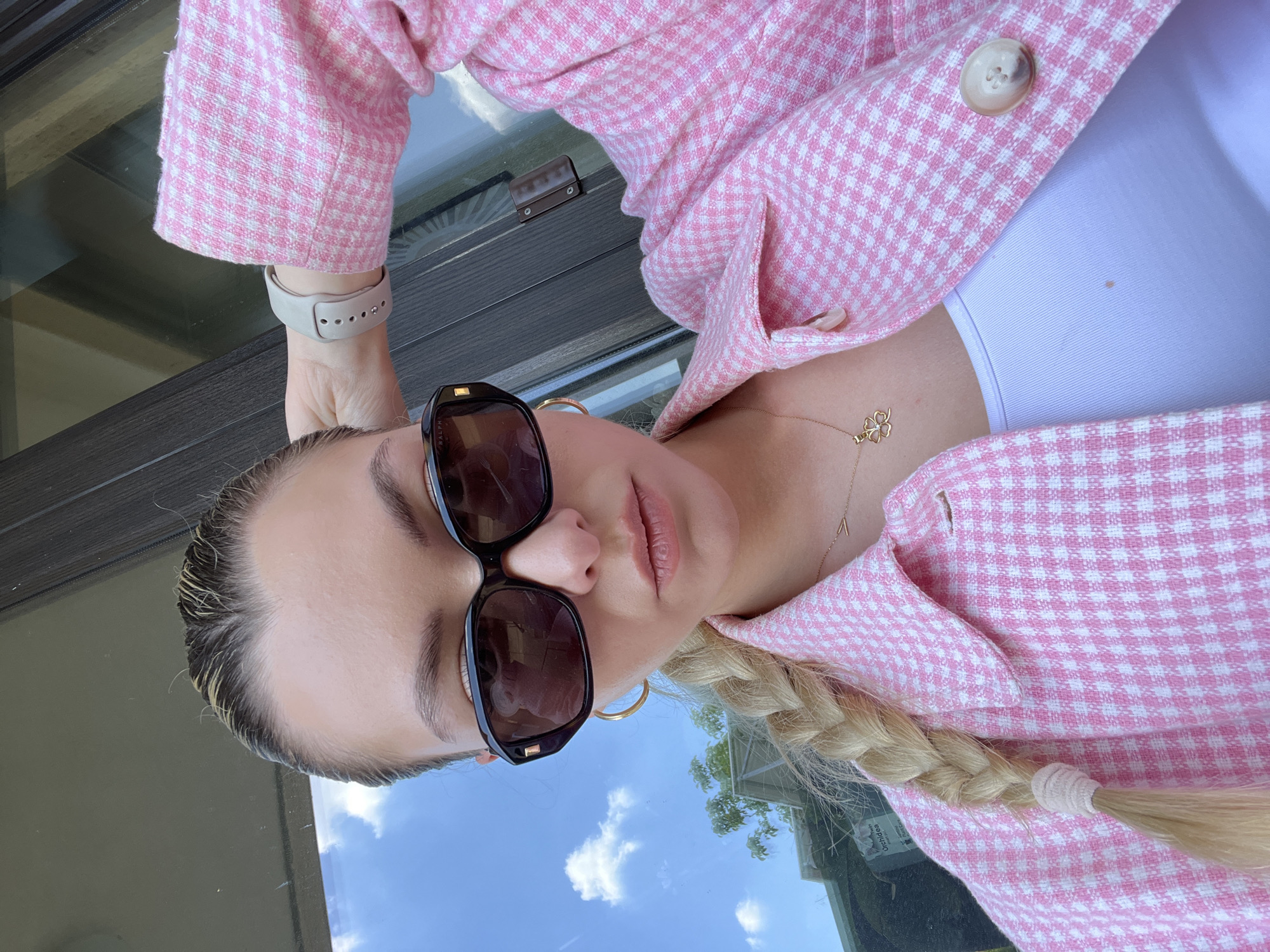 A woman is pictured from a medium-high angle in an indoor or semi-outdoor setting. She wears sunglasses, a pink and white checkered jacket over a white top, and has a light-colored braid. She has gold hoop earrings and a gold necklace with a clover-shaped pendant. She also wears a white smartwatch. The reflection in the sunglasses reveals a sky with scattered clouds, and a window or glass partition shows a building with greenery outside. The location is Unknown, Unknown.