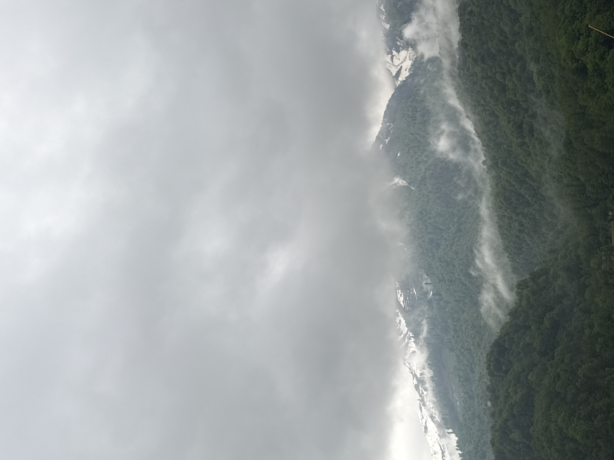 The image shows a mountain range in Krasnaya Polyana, Russia, partially obscured by heavy cloud cover. The mountains are covered in dense green forest, with patches of snow visible on the higher elevations. Mist or fog is also present, further obscuring the view. The sky is overcast and gray.