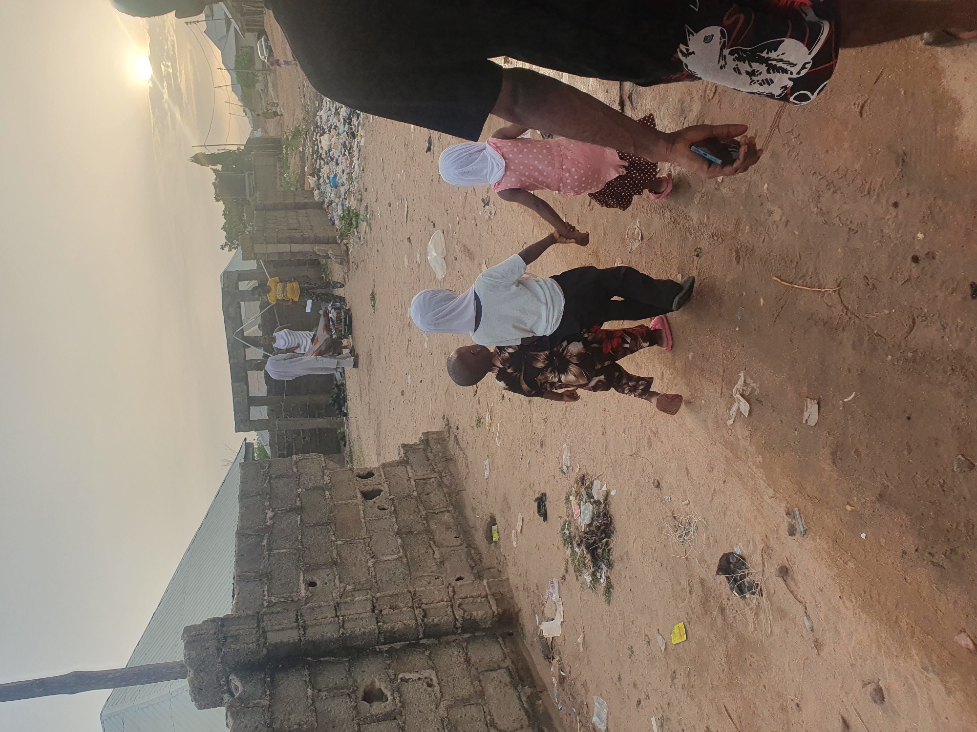 In Lokoja, Nigeria, a medium-angle shot captures a sandy, debris-strewn road. In the foreground, two children wearing white headscarves hold hands while walking. The first child wears a white top and black pants, while the second wears a pink dotted dress. An adult figure, partially visible in the foreground, is holding a smartphone. A partially constructed building stands nearby, with construction materials visible in the background. In the distance, three people are seen standing together. To the side of the road, there is a significant pile of trash. The sky is overcast, suggesting either early morning or late afternoon light.