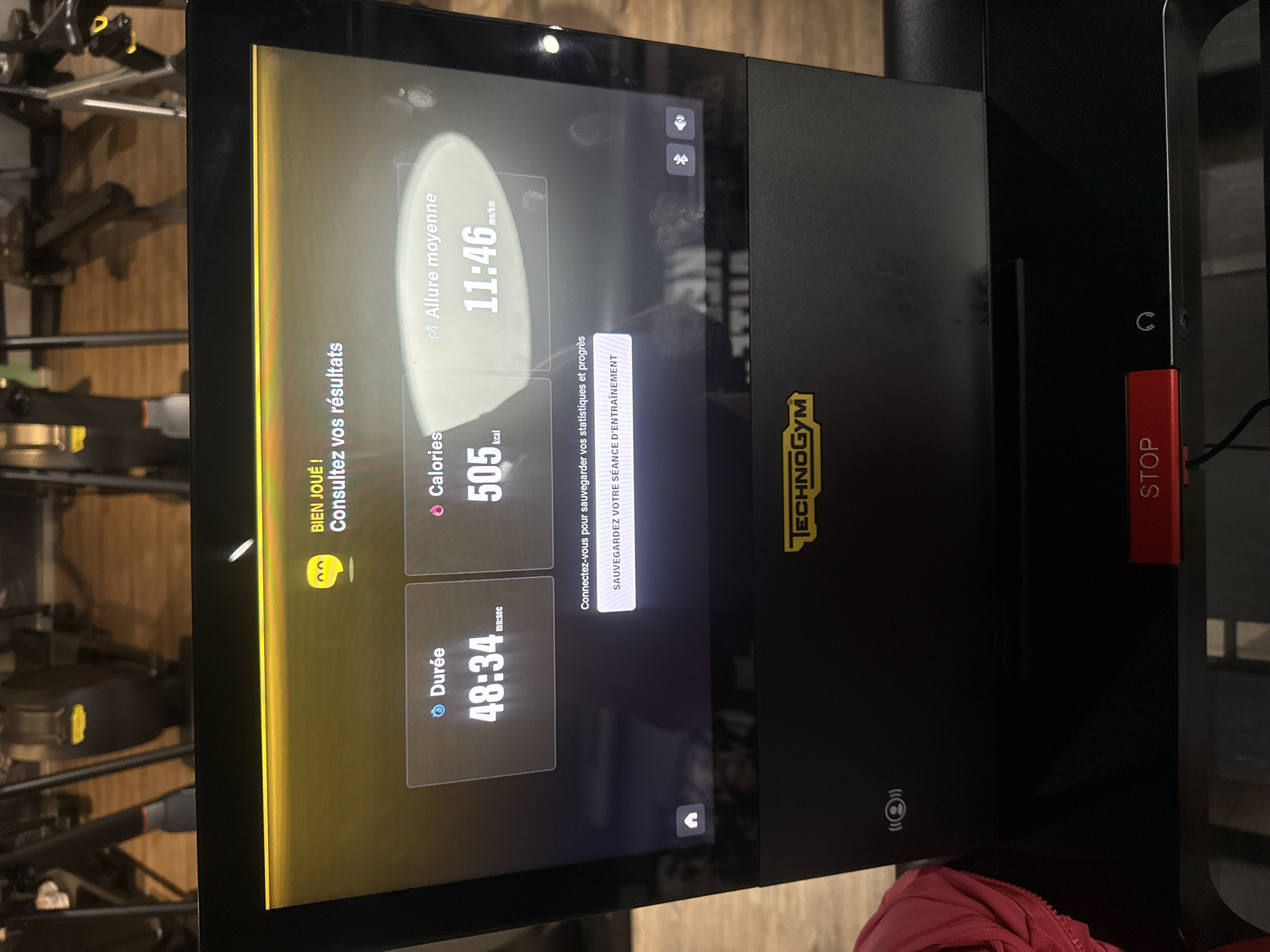 The image depicts the control panel of a Technogym exercise machine, possibly a treadmill. The display shows workout statistics: 48 minutes and 34 seconds of duration, 505 kcal burned, and an average pace of 11 minutes and 46 seconds per kilometer. The text "BIEN JOUE!" indicates that the workout has been completed. There is a prompt to save the workout session. The presence of the word "TECHNOGYM" indicates the brand of the equipment. A red "STOP" button and a headphone jack are visible on the right.
Given the French text and context, the image could be associated with a fitness facility in Paris, France.