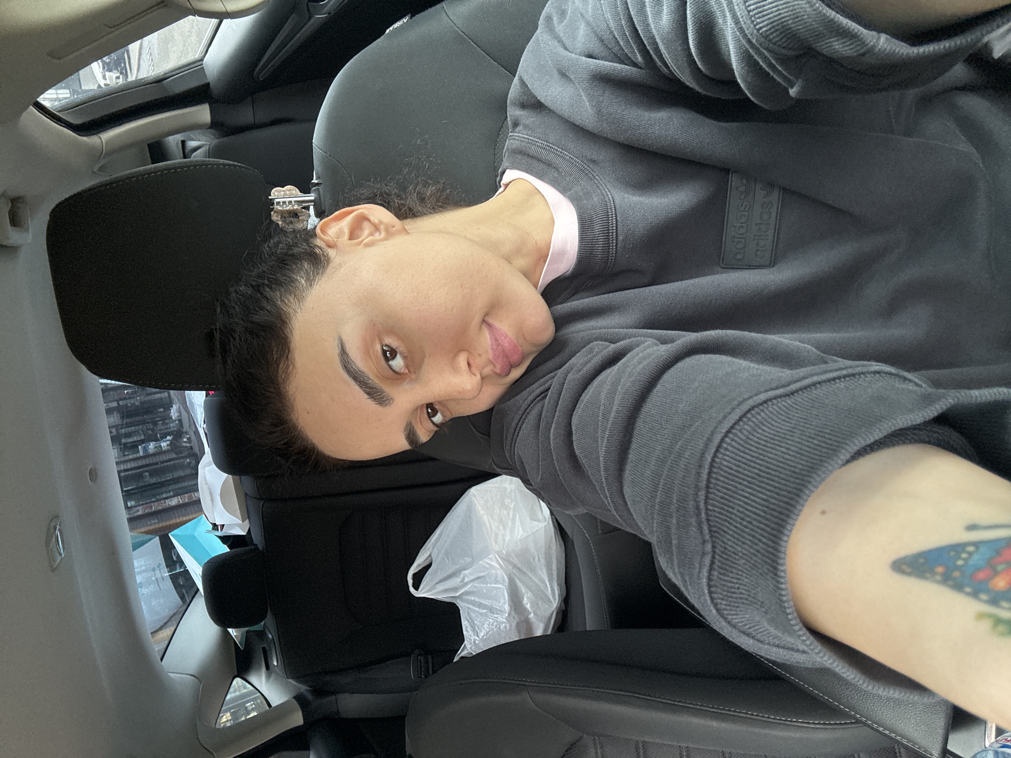 The image features a close-up selfie of a person inside a car. The person, with dark, curly hair pulled back, looks directly at the camera with a relaxed expression and a slight smile. They have prominent eyebrows and dark eyes. They are wearing a charcoal grey long-sleeved top, possibly a sweatshirt, with a light pink collar visible underneath. On the upper left sleeve of the grey top, a small, rectangular black patch is embossed with two vertically stacked "adidas" logos. A colorful tattoo, appearing to be a butterfly, is partially visible on their right forearm.
The setting is a car interior, characterized by dark seats and headrests. A white plastic bag rests on an adjacent car seat. A sunroof is visible in the upper part of the frame, and through the car windows, a blurred urban background with buildings and structures can be discerned, indicating a daytime, city environment. The word "AIRBAG" is faintly visible on an interior car pillar. The overall mood is casual. The provided context indicates the location is Tbilisi, Georgia.