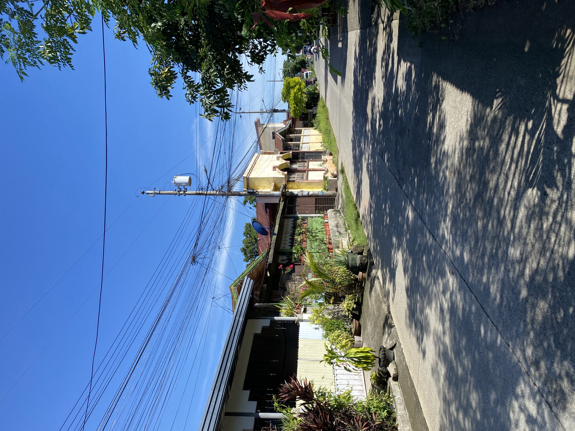 The image depicts a residential street in Davao City, Philippines, under a clear blue sky. A paved road runs diagonally from the foreground towards the background, lined with houses on either side. The houses appear to be single-story structures with various roof materials, including what looks like corrugated metal and asphalt shingles. Many of the houses have plants and potted greenery along their facades, suggesting a well-maintained residential area. Overhead, a dense network of electrical wires crisscrosses the sky, supported by utility poles. One pole, equipped with a transformer, is clearly visible. Sunlight casts dappled shadows from the foliage onto the road, indicating a bright, sunny day. In the distance, a few vehicles and a motorcycle can be seen, suggesting normal street activity. The overall impression is a typical urban residential street scene in a tropical climate.