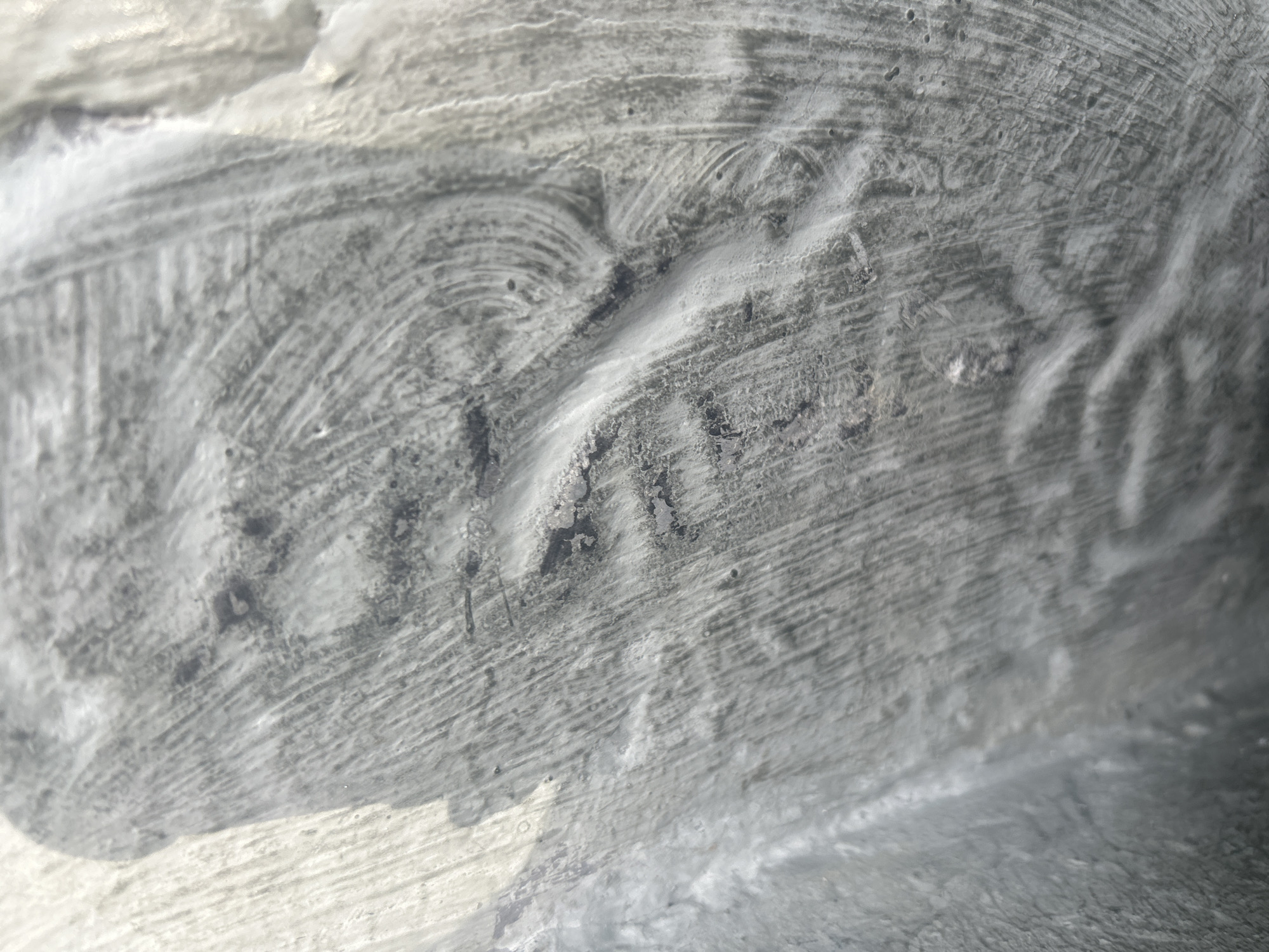 The image displays a close-up, abstract view of a rough, textured surface, predominantly gray and white in color. The surface appears to be a wall or a similar structural element with visible brush strokes, imperfections, and layered paint or plaster. There are discernible patterns of curved and linear marks, suggesting application by hand or with a tool. The lighting creates shadows and highlights that emphasize the unevenness and topography of the surface. There are no people or identifiable objects present in the frame. The environment is indeterminate and does not provide any specific clues to its location within Santa Ana, Venezuela, or any other geographical setting.