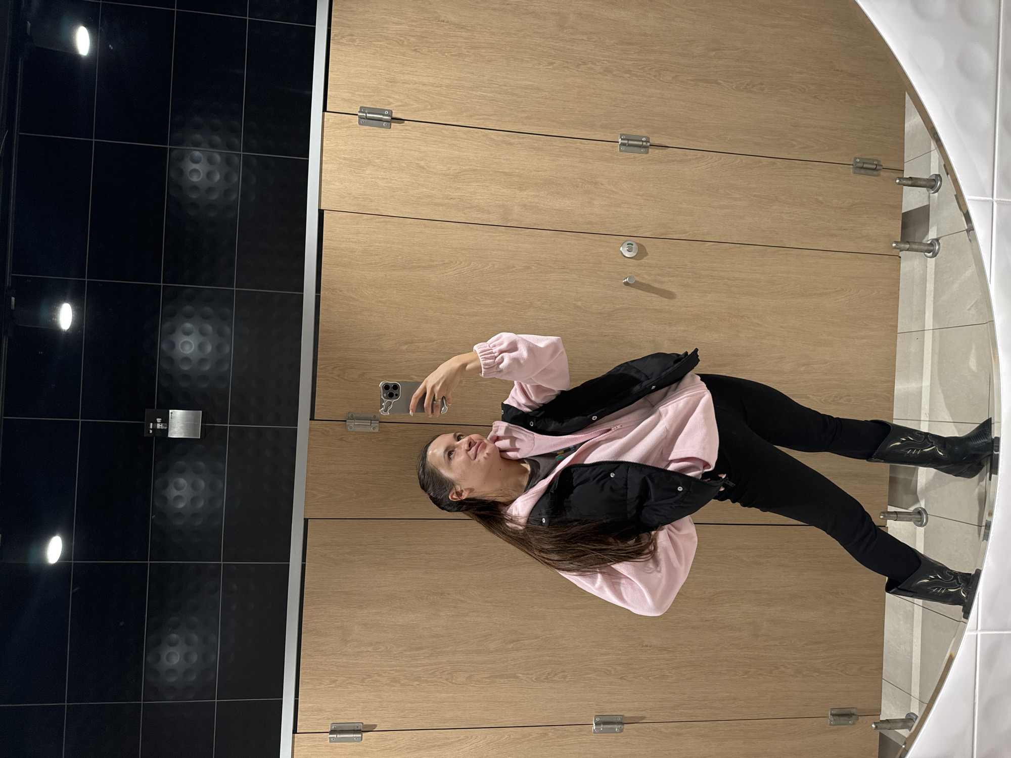 A woman is taking a selfie in a restroom. She is wearing black pants and cowboy boots, a pink hooded sweatshirt, and a black puffer vest. The restroom has light-colored wooden stall doors, black tiled walls, and a mirror. It is unknown whether the location is in Ivashevo, Russia.