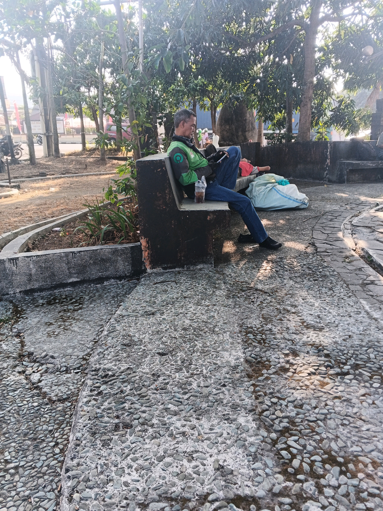 In the city of Ungaran, Indonesia, a man in a green jacket with a green circular logo sits on a concrete bench in an outdoor park. He is looking down at his phone, which he holds in his left hand. A plastic cup with a straw sits on the bench next to him. To his right, another person is lying on the ground, covered by a light blue bag. The ground is paved with small, gray stones. Trees with lush green leaves are visible in the background, and the sun is shining, casting shadows on the ground.
