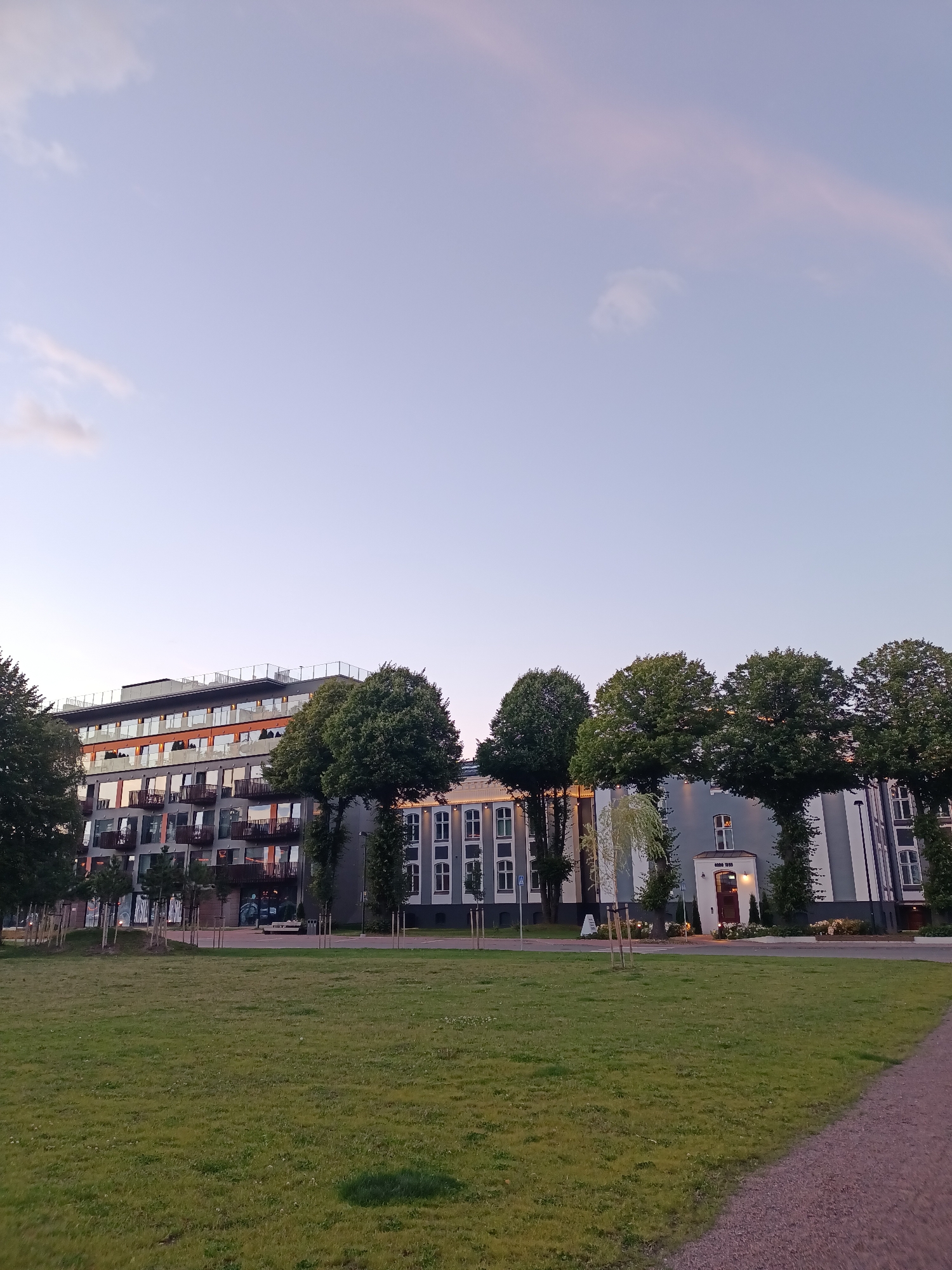 The image shows a modern apartment building with balconies and large windows, reflecting the twilight sky. In front of the building is a line of mature trees, and beyond them, a well-maintained grassy lawn slopes down to a gravel path. The sky is a soft gradient of pale blue and pink, indicating it is either sunrise or sunset. The apartment building features a mix of grey and orange facades, with a contemporary design. To the right of the apartment building, a more traditional, lighter colored building with arched windows is visible. This scene is located in the Kopli district of Tallinn, Estonia.