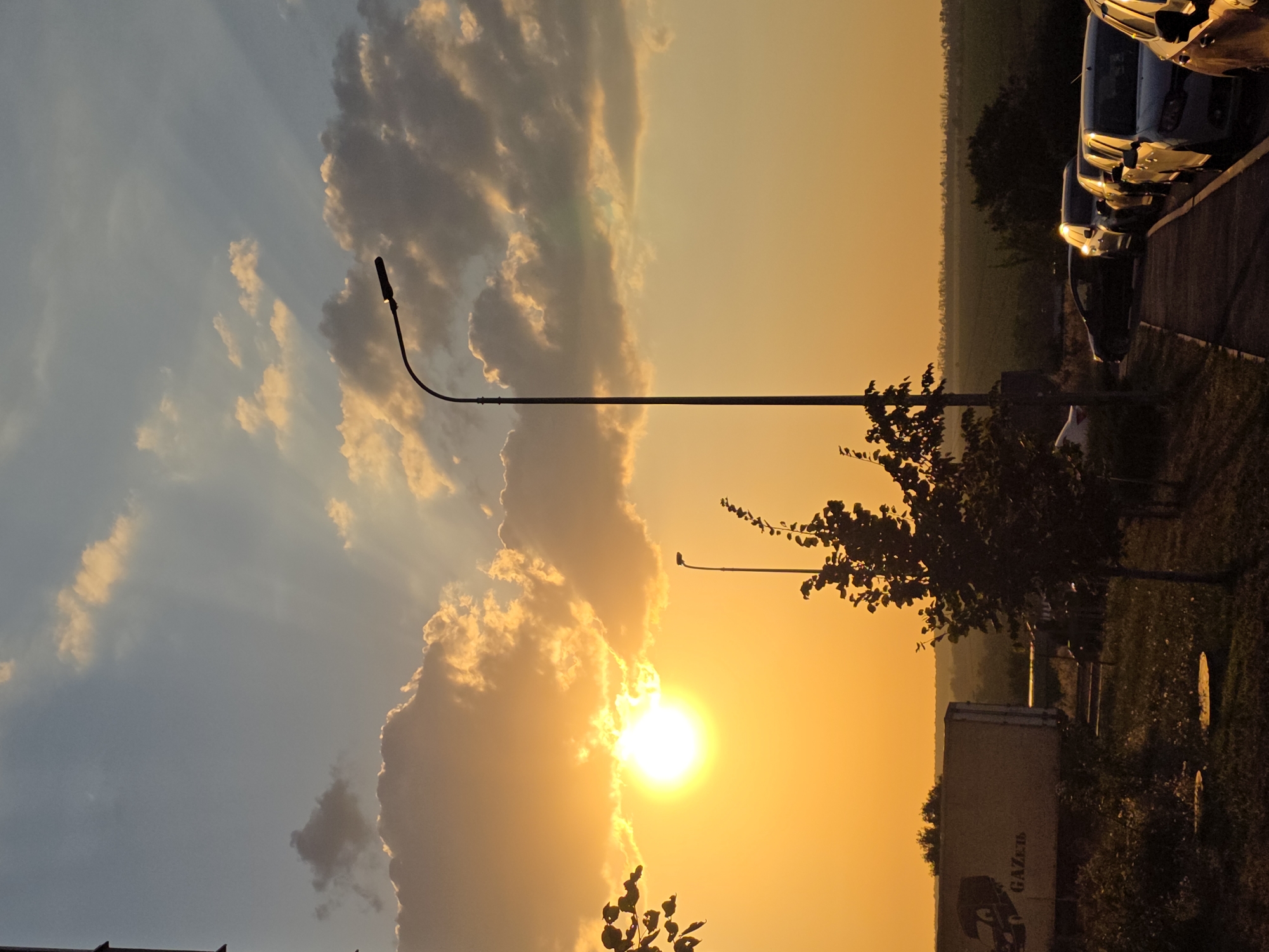 The image depicts a sunset scene in Rostov-on-Don, Russia. The sun, a bright orb of light, is low in the sky, casting a warm, golden glow across the scene. Rays of sunlight pierce through scattered clouds, creating dramatic light effects. In the foreground, a streetlight stands tall on the right side of the frame. A line of parked cars is visible on the right, reflecting the warm light of the sunset. On the left, a white truck with the word "GAZel" printed on its side is partially visible. Trees and foliage are present in the midground and foreground, with some appearing silhouetted against the bright sky. The background shows a hazy horizon, suggesting fields or rural landscape. The overall impression is a peaceful, serene evening in the city.
