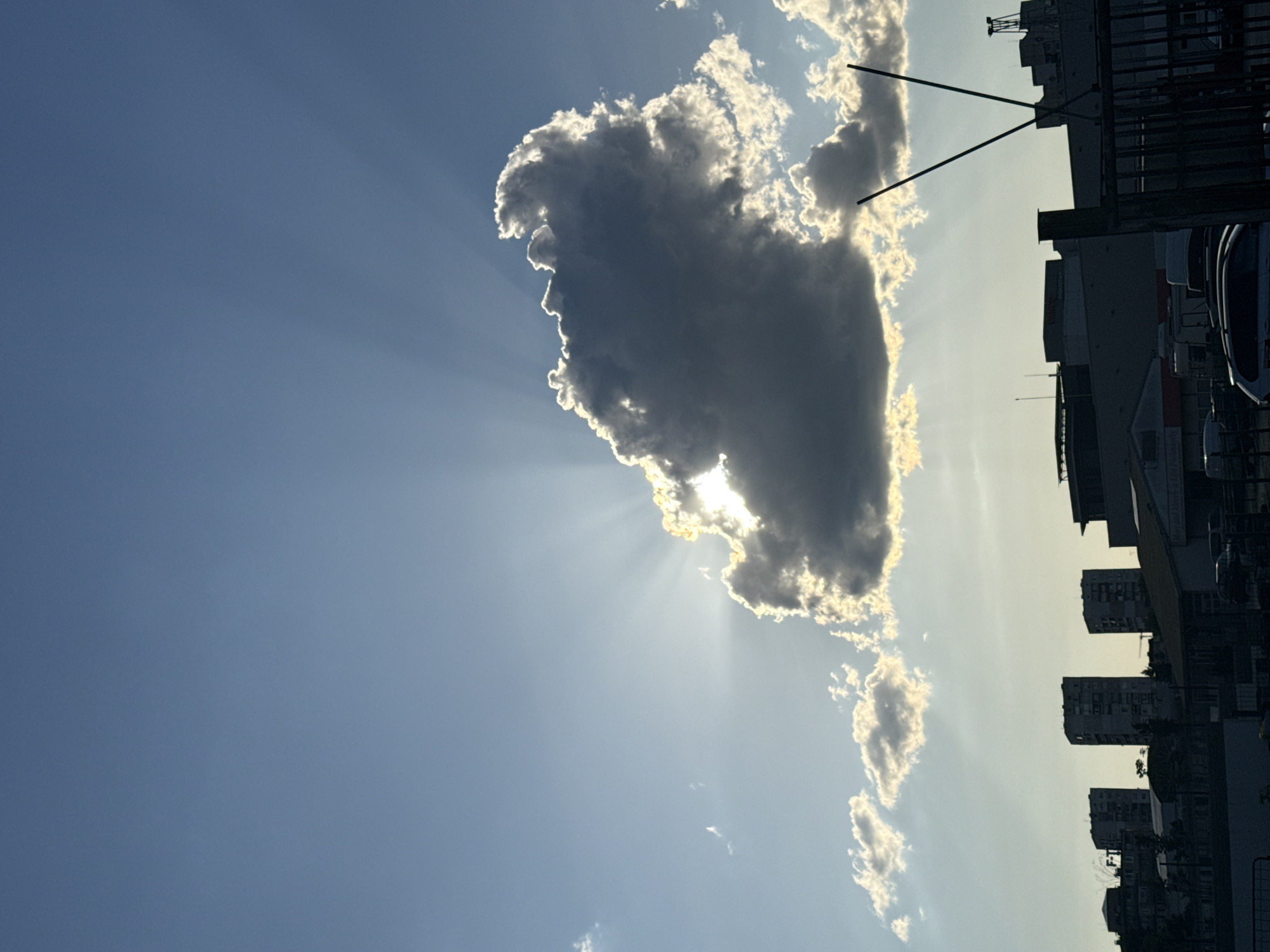 A low-angle, rotated, sunlit, daytime shot shows a cloud with sunrays protruding from it, a clear blue sky, and buildings silhouetted against the bright sky. The location is identified as Sofia, Bulgaria.