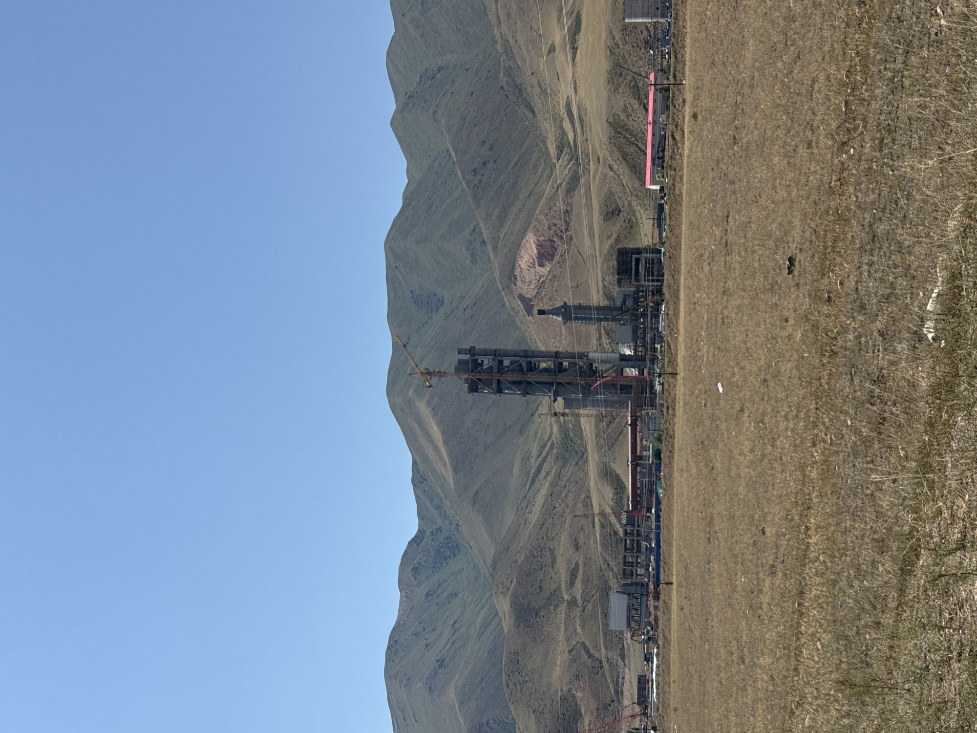 The image depicts an industrial construction site in the city of Sasyk-Bulak, Kyrgyzstan. In the foreground, a dry, grassy field stretches towards the construction zone. The construction area features several large, unfinished structures, including a tall, multi-story tower crane and a series of interconnected buildings. Red and yellow cranes are visible amidst the construction. The background is dominated by rolling, barren hills under a clear blue sky. The scene conveys a sense of ongoing development in a semi-arid, mountainous environment.
