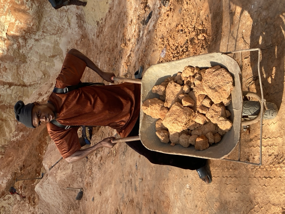 A person stands next to a wheelbarrow filled with dirt and rocks, possibly in a mining or quarry setting. The individual wears a brown shirt and a black hat. They appear to be holding the handles of the wheelbarrow, ready to move the load. The background shows a dirt wall or hill, suggesting an excavated area. A few tools such as a shovel are visible nearby.