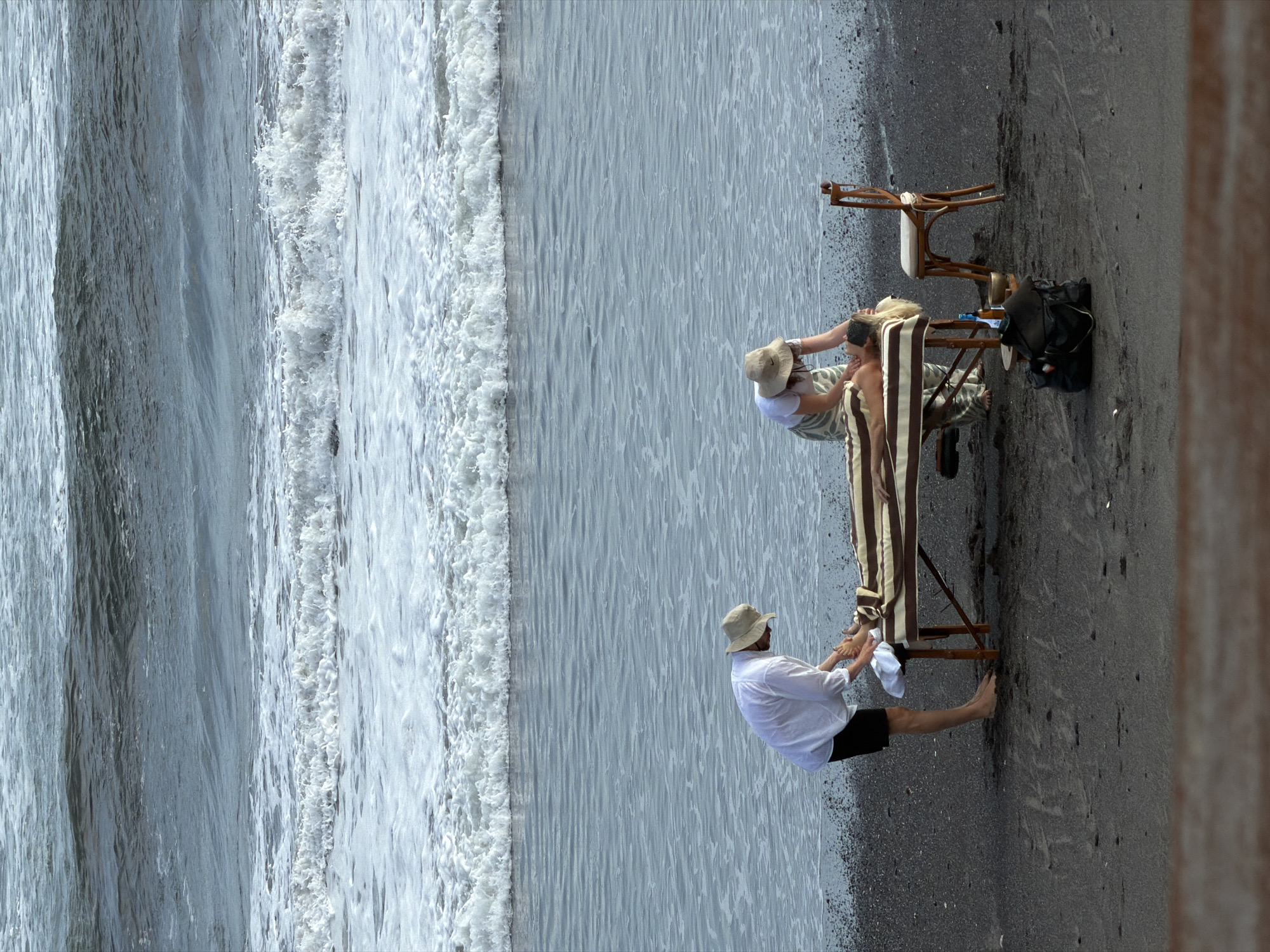 Two people are on a beach next to ocean waves. One person lies on a striped chaise lounge, wearing a white hat. The other person is sitting and attending to the person on the lounge chair. A chair, bag, and other items are next to the lounge chair. The beach is black.