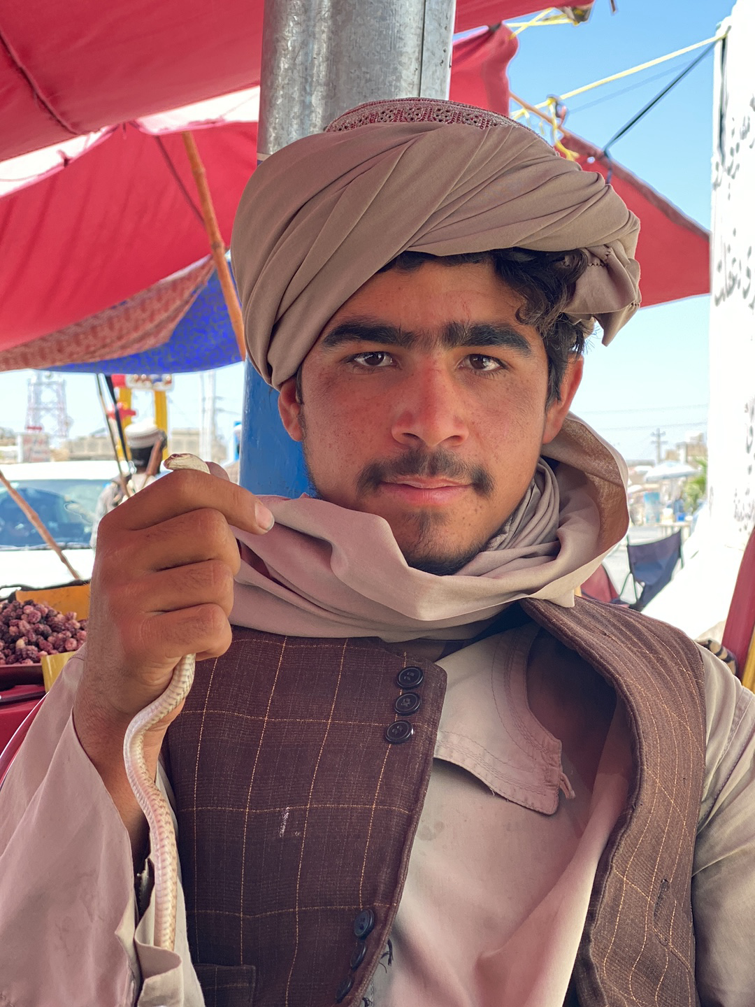 A medium shot depicts a man holding a snake. He wears a tan turban and a brown, grid-patterned vest over a lighter-colored shirt. A tan scarf is draped around his neck. The man is facing the camera with a neutral expression. In his left hand, he holds a pale snake, possibly albino. The background reveals a bustling outdoor market scene with red and blue canopies. Other people and possibly fruits and vegetables are partially visible. A vehicle is seen in the distance. The image is taken in daylight and the environment suggests قندهار, Afghanistan.