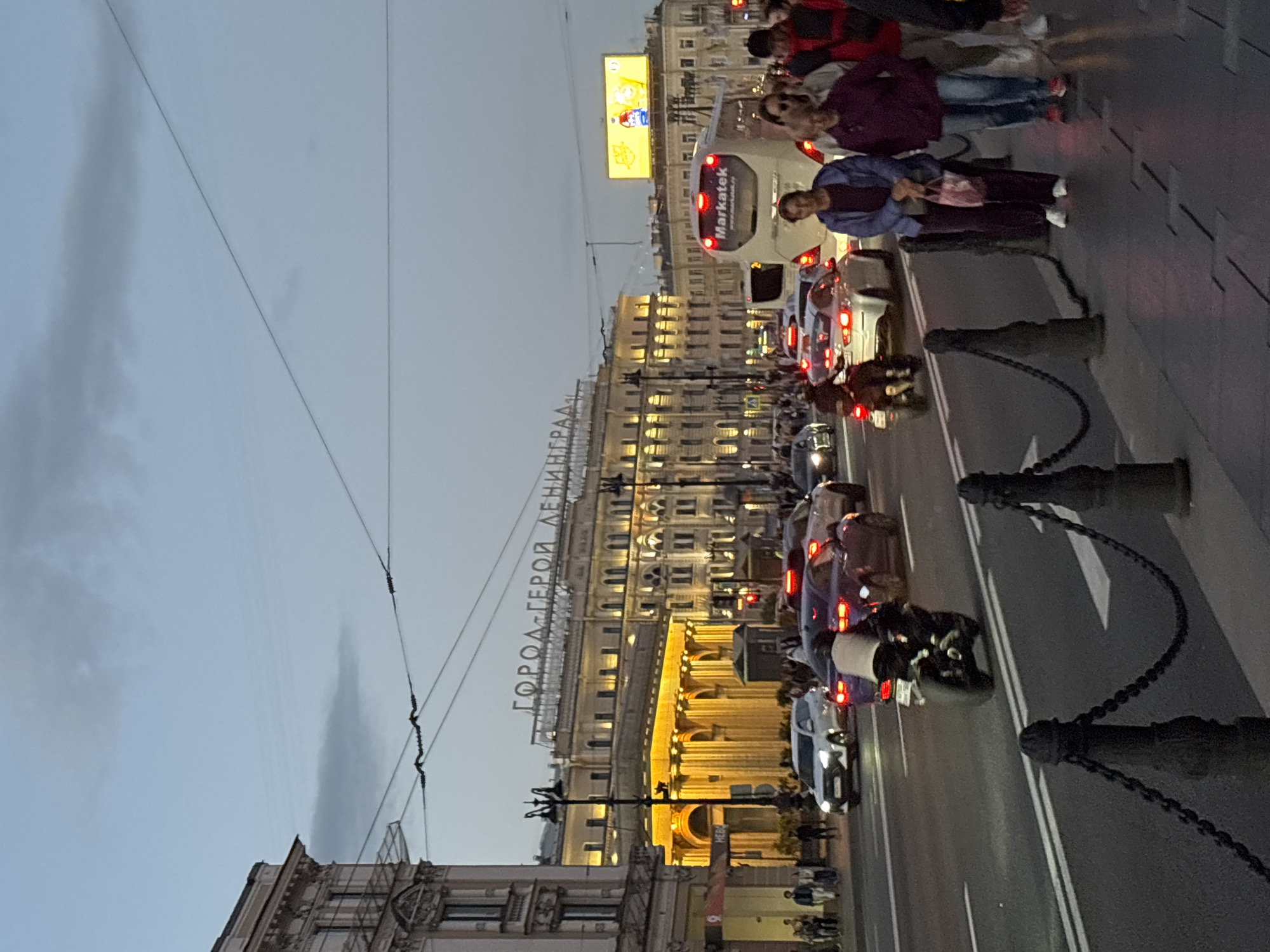 The image shows a street scene in Saint Petersburg, Russia, likely on Nevsky Prospekt, a major avenue. The time appears to be dusk or early evening, with the sky a soft grey and the city lights beginning to illuminate the buildings.
In the background, a large, ornate building dominates the skyline. Above this building, a sign in Russian reads "ГОРОД-ГЕРОЙ ЛЕНИНГРАД" (City-Hero Leningrad), a historical designation for Saint Petersburg during World War II. The building itself has numerous windows and architectural details, with warm yellow light emanating from some sections, suggesting businesses or public spaces within.
The street is bustling with activity. Several cars are visible, including a white SUV and a dark-colored sedan. A motorcycle with a rider is in motion on the road. A white bus with "Markatek" branding is also present, indicating public transportation or a commercial service.
On the sidewalk, a group of people are walking. They are dressed in casual clothing, and some are carrying bags. The sidewalk is paved with stone, and a decorative metal fence with chains lines the edge of the road. Streetlights are also visible, casting a dim glow on the scene. The overall impression is of a lively urban environment at the transition from day to night.
