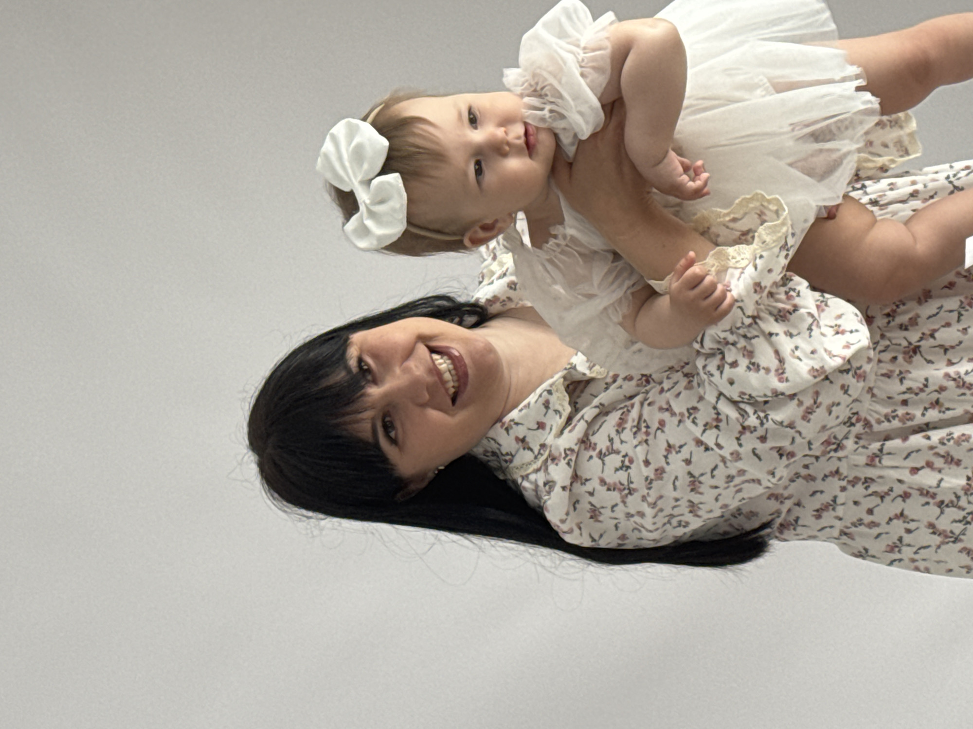 A woman with black hair is holding a baby girl in a white dress with a large white bow in her hair. Both are smiling. The background is plain and light-colored. The location is not identifiable from the image.