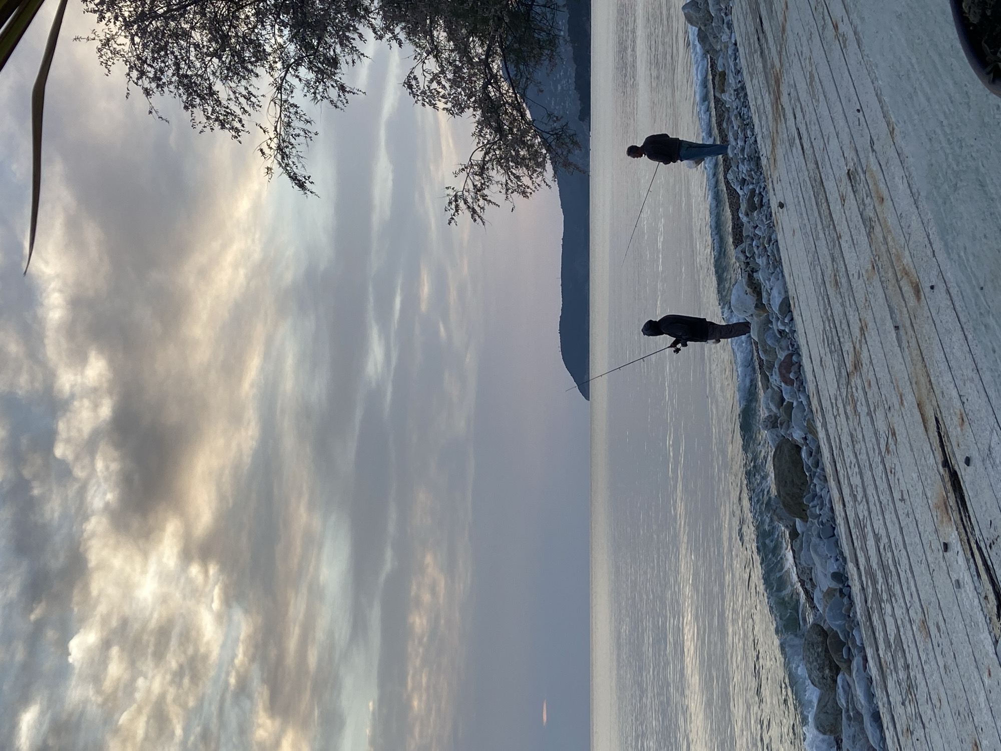 Two people are fishing on a wooden pier in Bar, Montenegro. The sky is cloudy and a hill is visible in the distance.