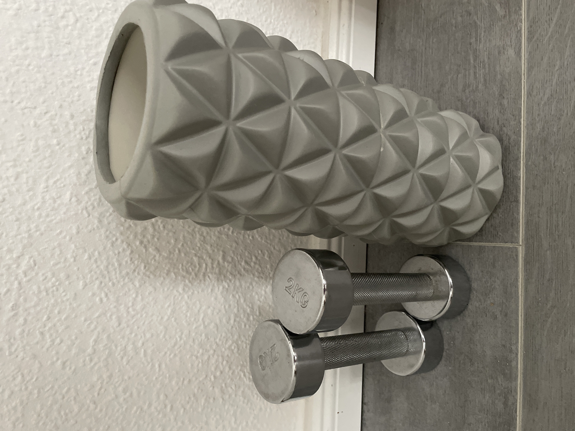 Two silver dumbbells are placed on a gray tiled floor in front of a textured white wall. To the right of the dumbbells is a tall, gray, geometric-patterned vase. The dumbbells appear to be 2kg each, as indicated by the markings on their tops. The scene is indoors, with the floor tiles and wall suggesting a domestic setting.