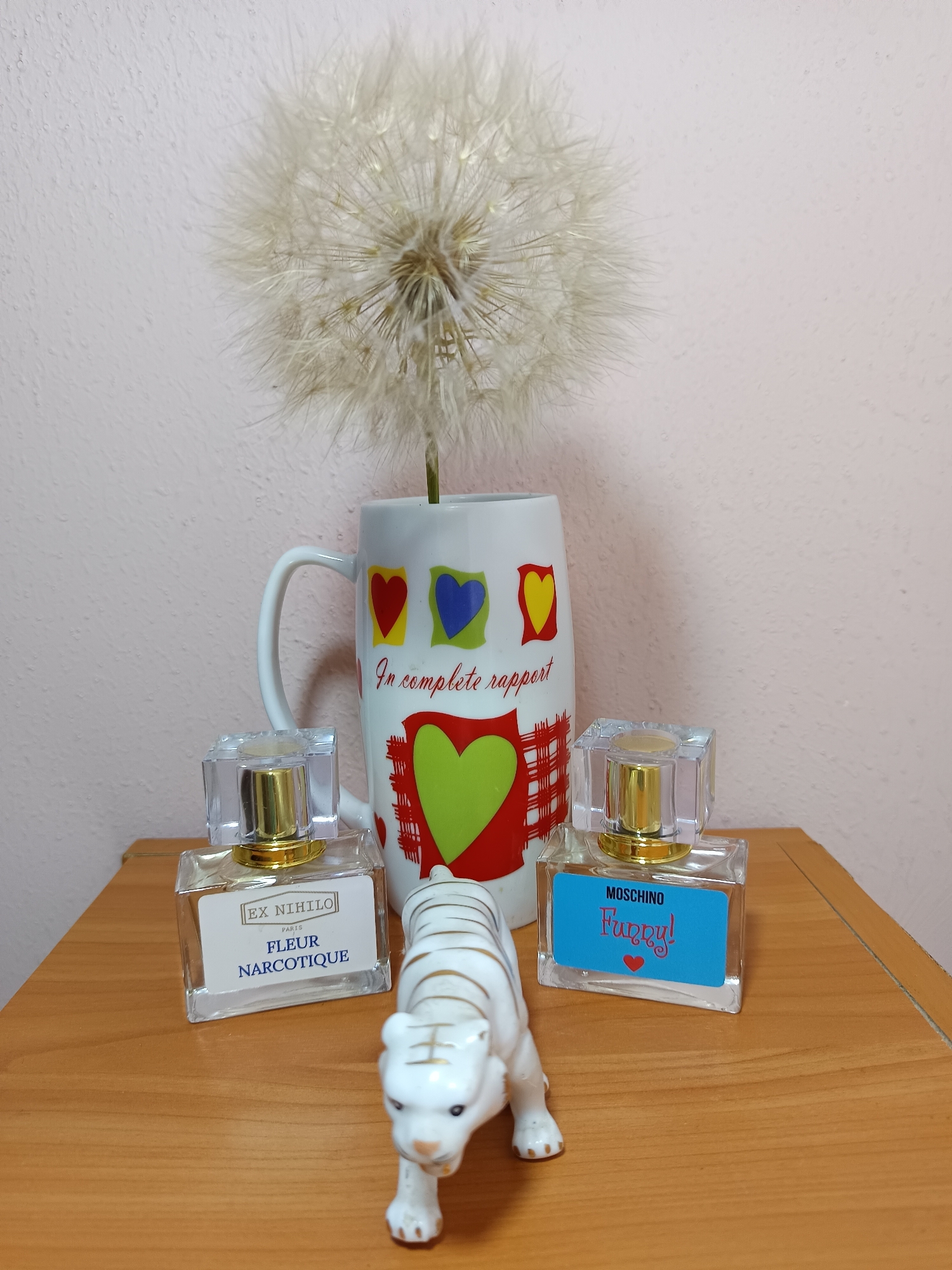 A still life arrangement sits on a wooden table against a light pink textured wall. In the center of the image, a white ceramic mug with a handle is positioned vertically. The mug is decorated with various colored hearts: a red heart, a yellow heart, and a blue heart near the top, and a large green heart with red criss-cross lines above it towards the bottom. A large dandelion seed head is placed inside the mug, with its fluffy white filaments extending upwards.
To the left of the mug, a clear glass perfume bottle with a gold cap sits on the table. A white label on the bottle reads "EX NIHILO PARIS FLEUR NARCOTIQUE". To the right of the mug, another clear glass perfume bottle with a gold cap is present. This bottle has a bright blue label that reads "MOSCHINO Funny!" in red cursive lettering, with a small red heart below the text.
In front of the perfume bottles, a small, white ceramic figurine of a tiger with gold stripes is positioned, facing slightly towards the viewer. The tiger appears to be walking. The wooden table surface is visible in the foreground, with its grain patterns apparent. The background wall has a subtle, uneven texture and a light pink hue. The image appears to be taken indoors.